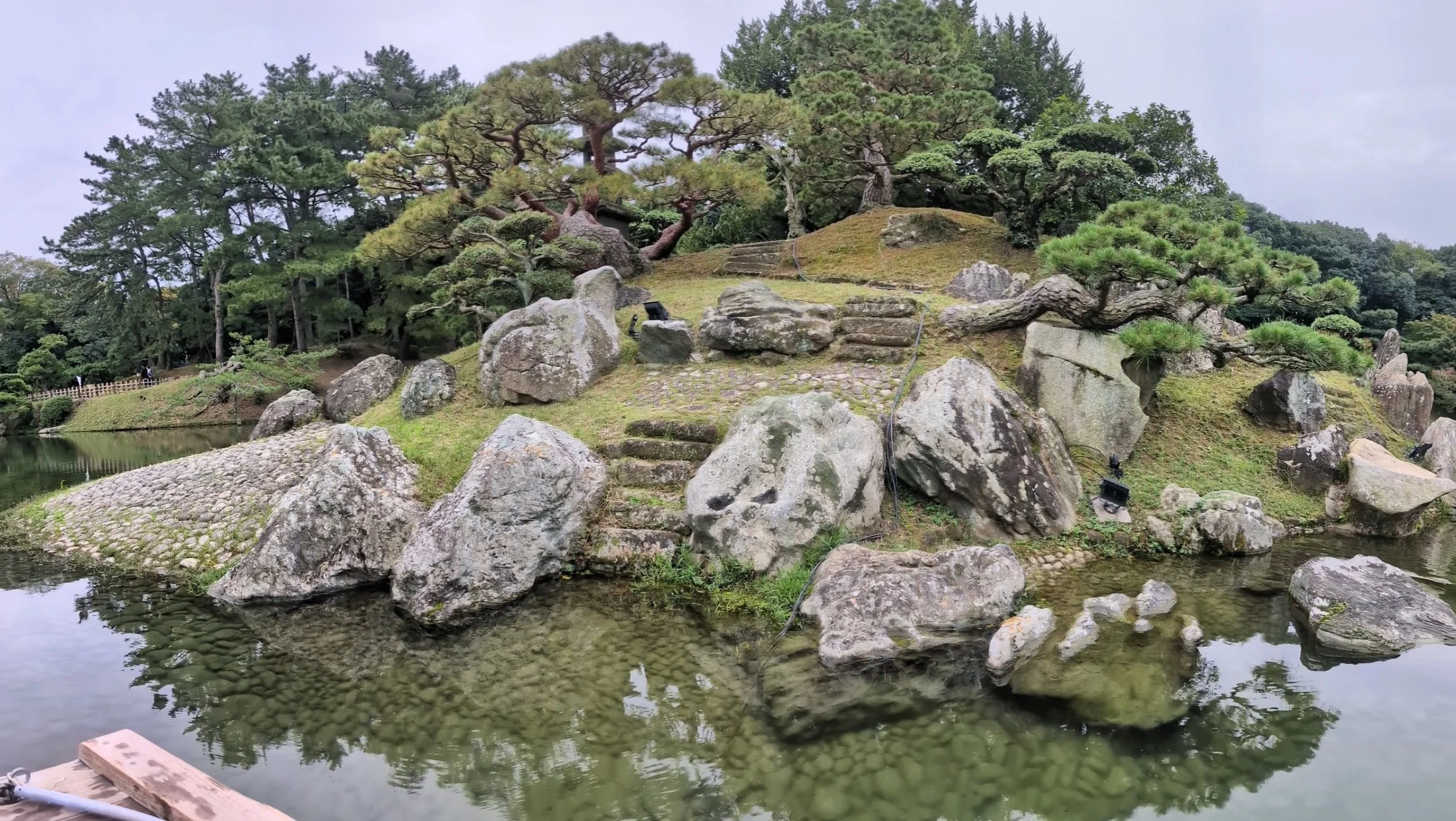 A traditional Japanese garden with a small pond, rocks, and pruned trees, including a pine, on a cloudy day.