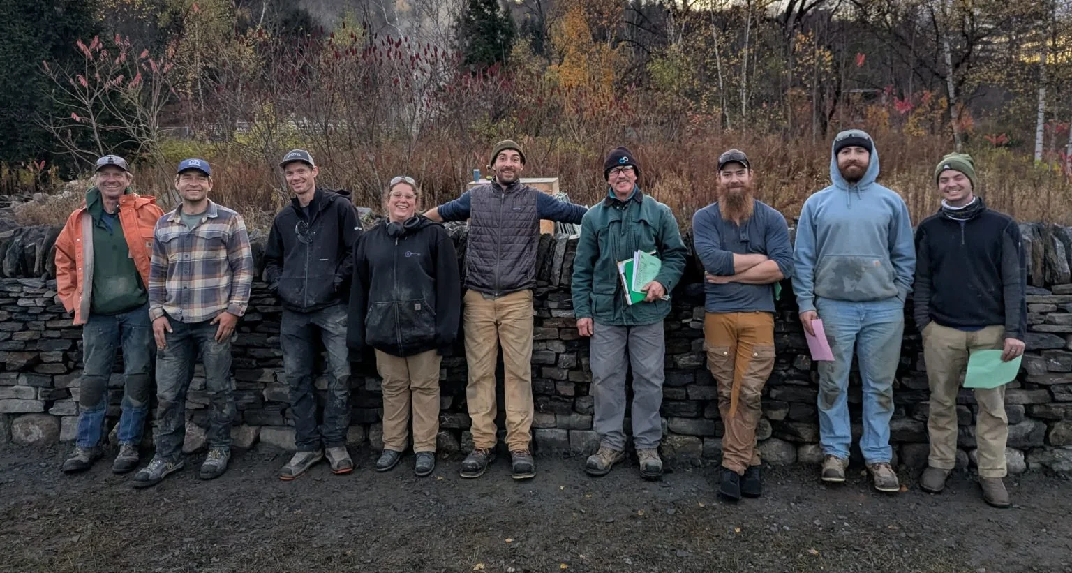 Group of nine people standing outdoors in front of a stone wall, smiling and posing for the photo, with autumn trees in the background.