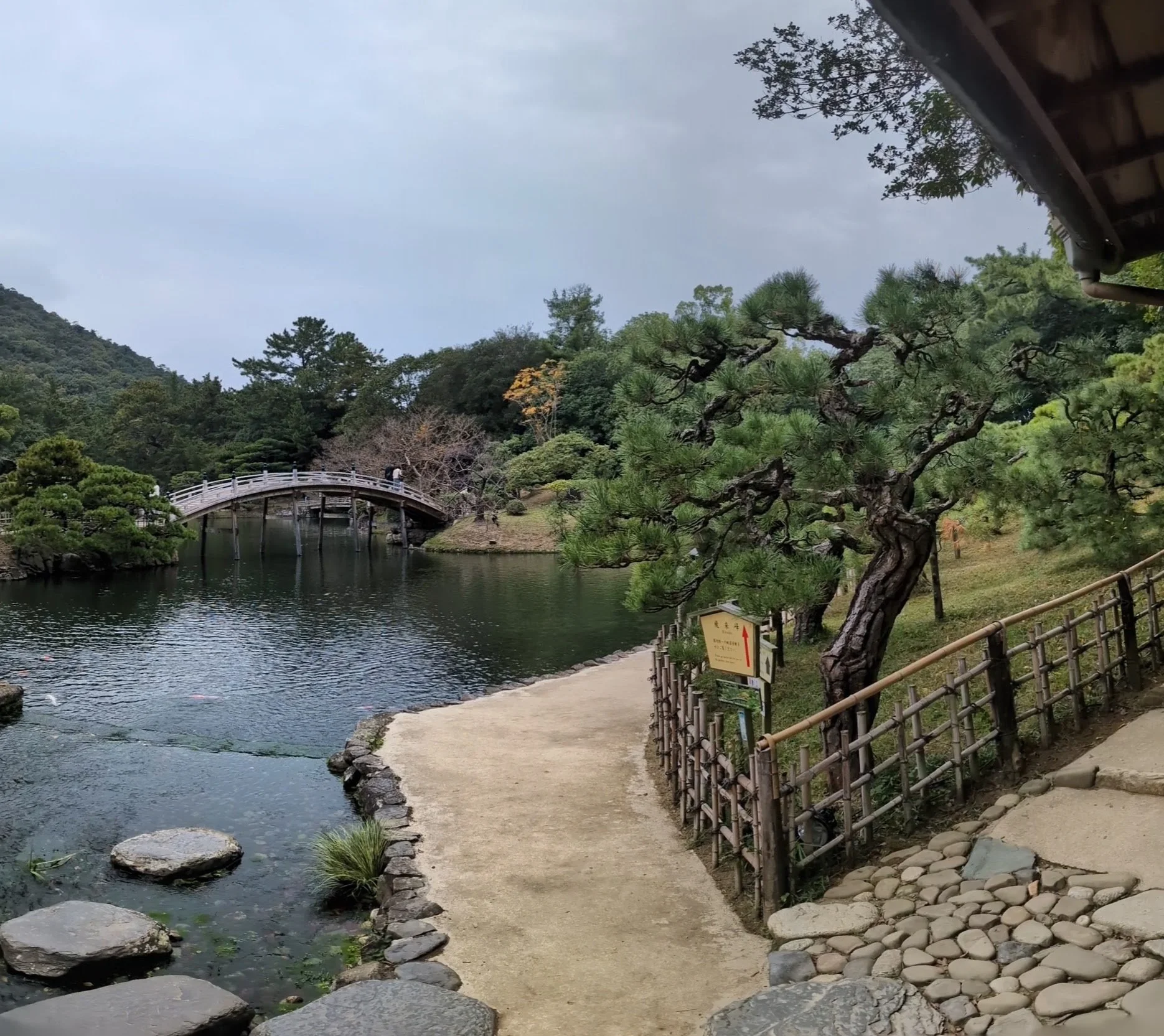 A peaceful Japanese garden with a small bridge over a pond, surrounded by trees and a stone pathway with a wooden fence.