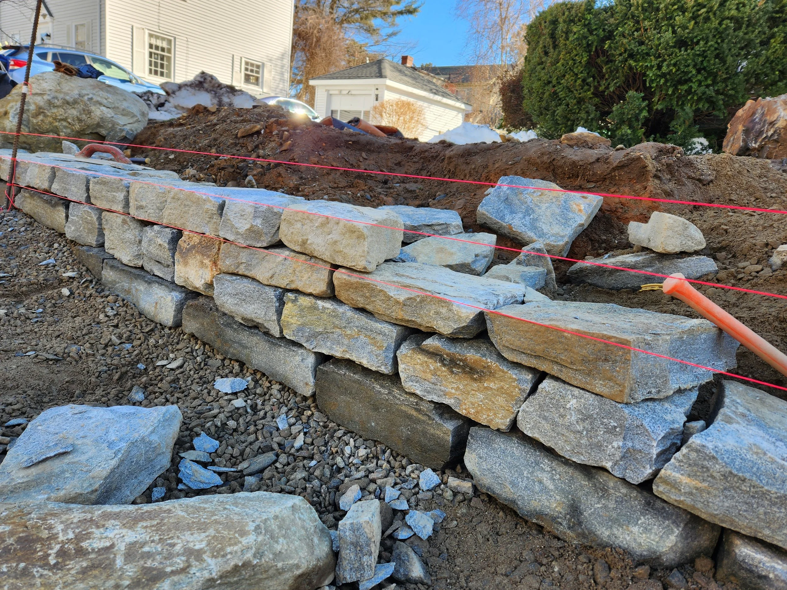 Construction site with a partially built stone wall, orange string line for alignment, and scattered rocks and dirt around.