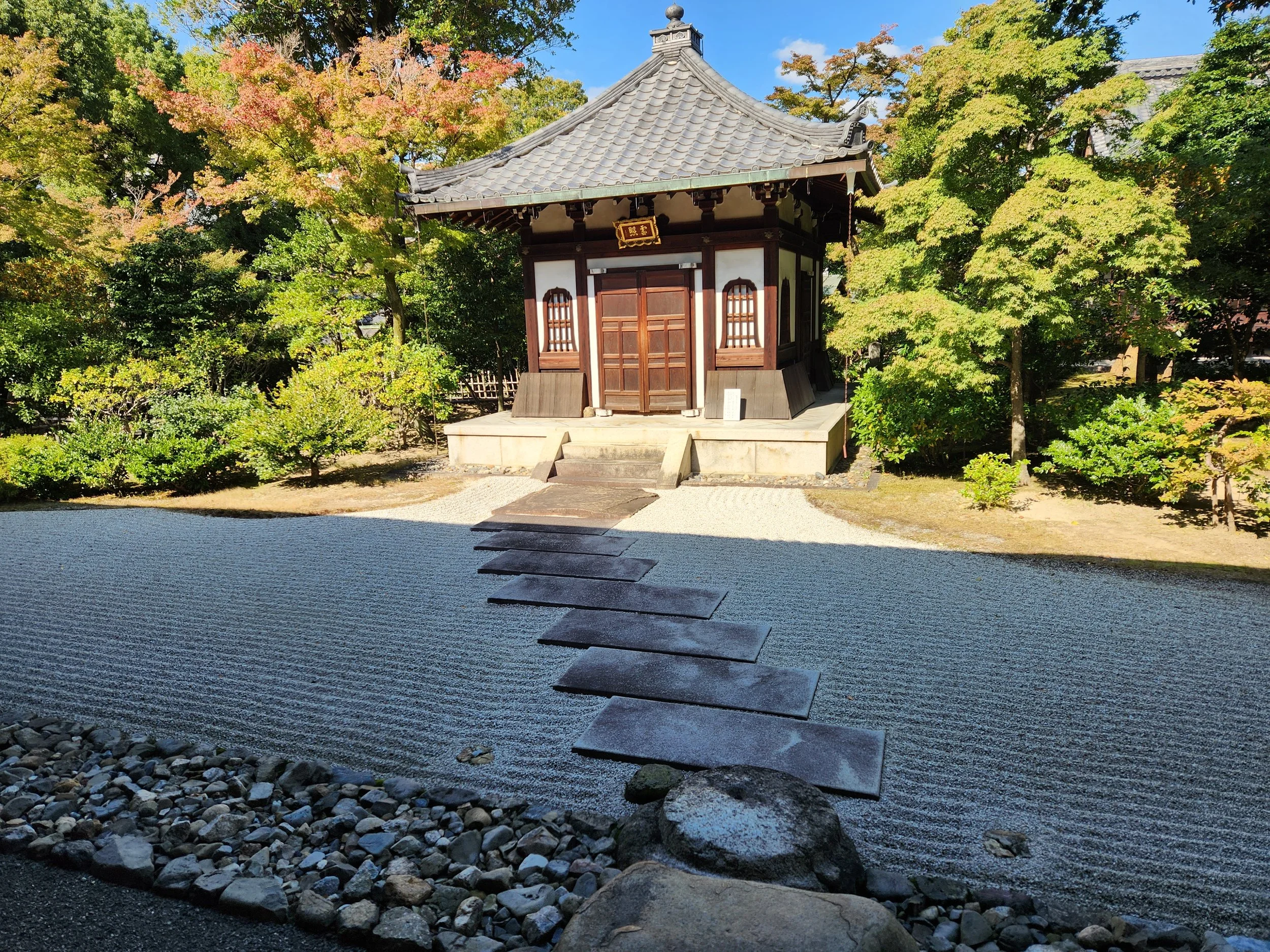 A traditional Japanese garden with a small wooden structure in the center, surrounded by lush green trees. There is a stepping stone pathway leading to the structure and a neatly raked gravel area.