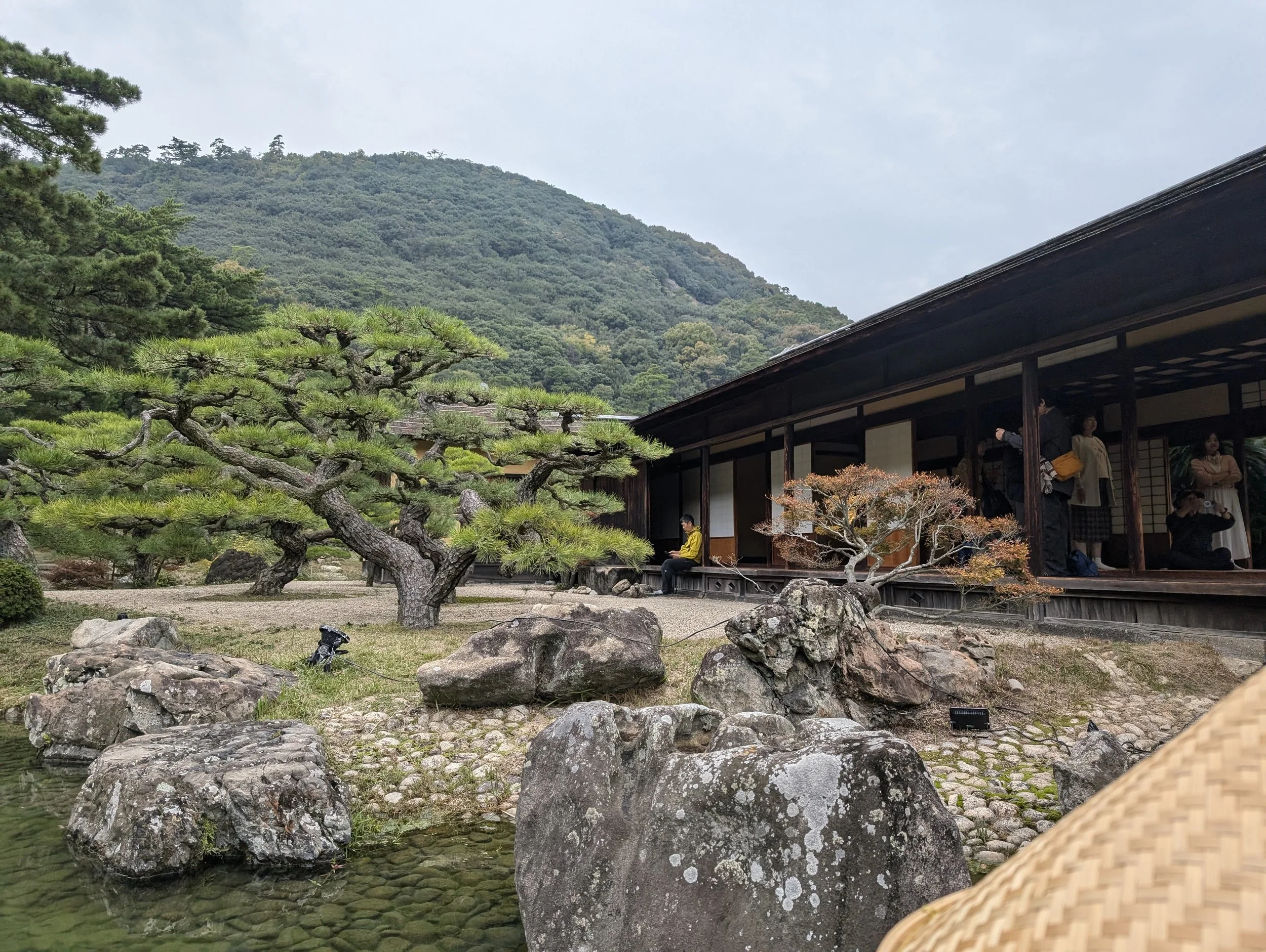 Traditional Japanese garden with pruned pine trees, rocks, and a pond in the foreground. A wooden building with sliding doors is visible on the right, with several people inside and one person sitting on a bench outside. In the background, a mountain