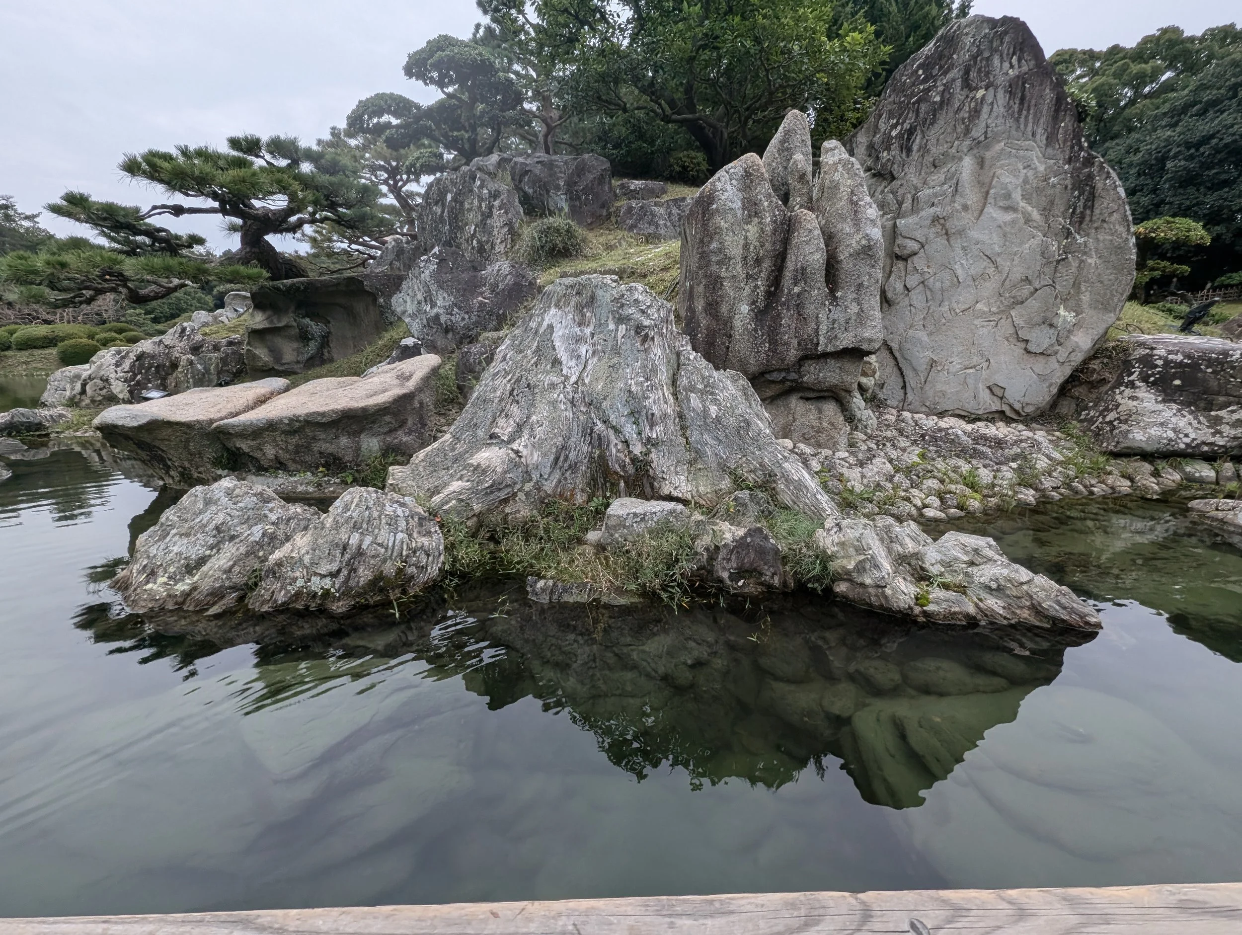 A traditional Japanese garden features large rocks and a pond, with trees and greenery in the background.