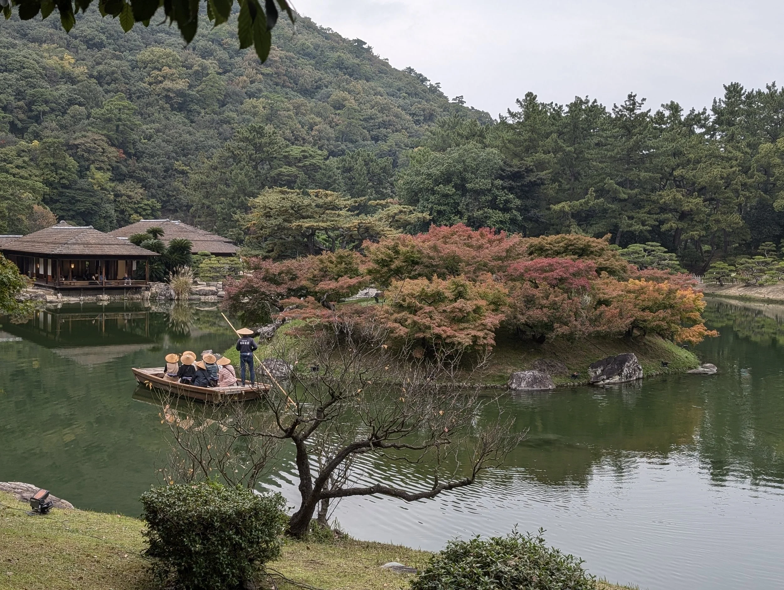 A traditional Japanese garden with a pond surrounded by lush trees, including a large pink and orange maple tree. A small boat with four tourists wearing straw hats is being paddled by a guide. Traditional buildings are visible in the background.