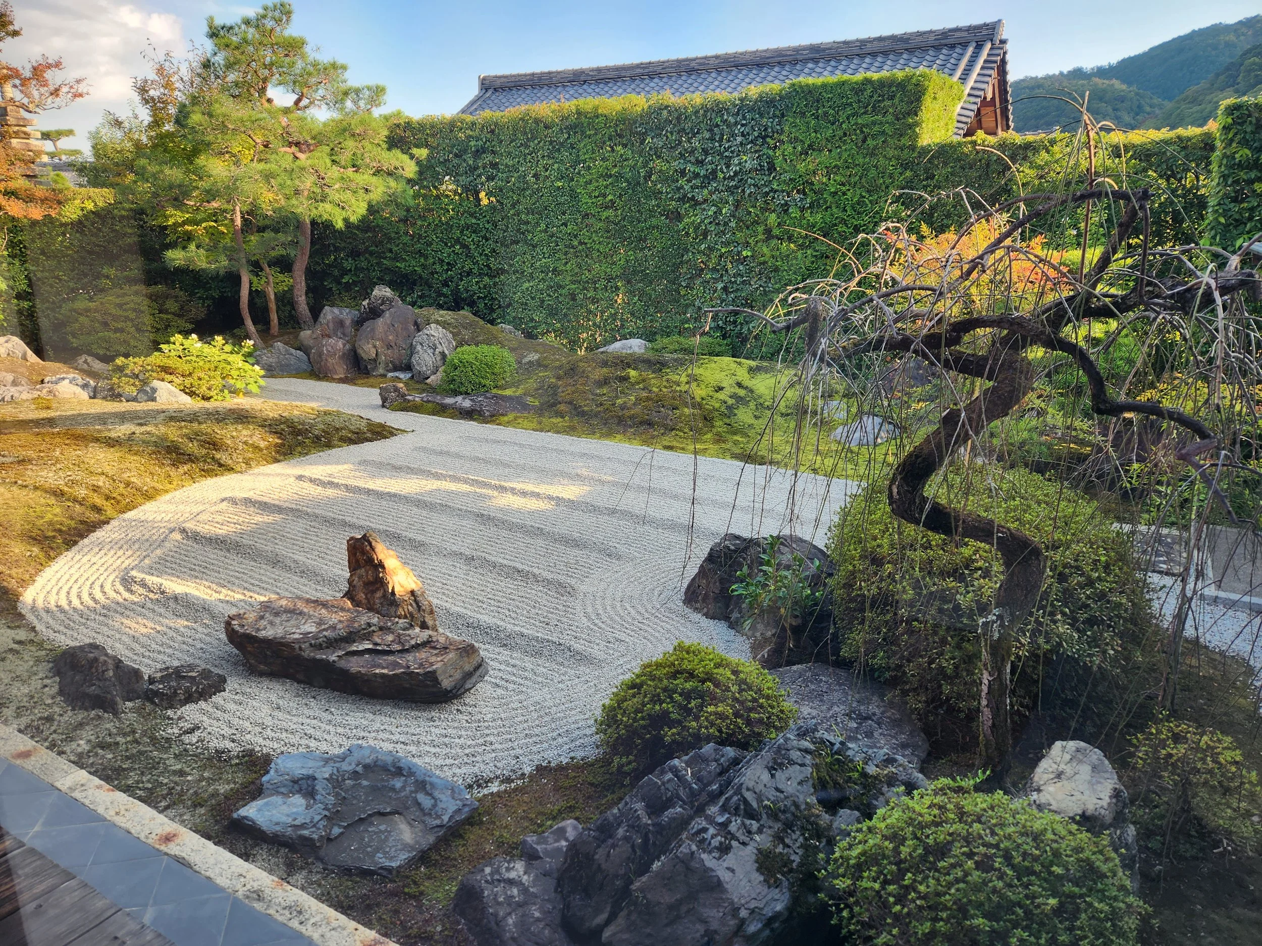 Japanese Zen garden with raked gravel, rocks, moss, small trees, and a traditional building in the background.