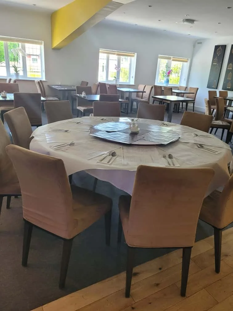 Empty restaurant dining area with round table set with utensils, beige chairs, and windows with natural light.