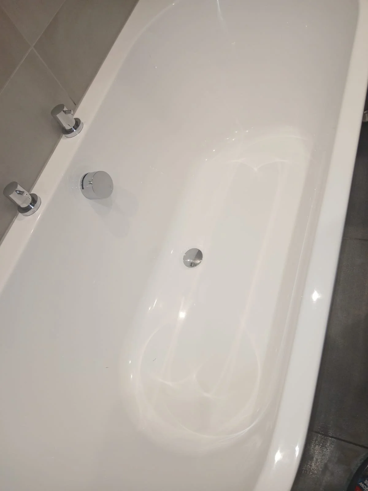 Empty white bathtub with silver faucet and drain, bathroom wall with gray tile on one side.