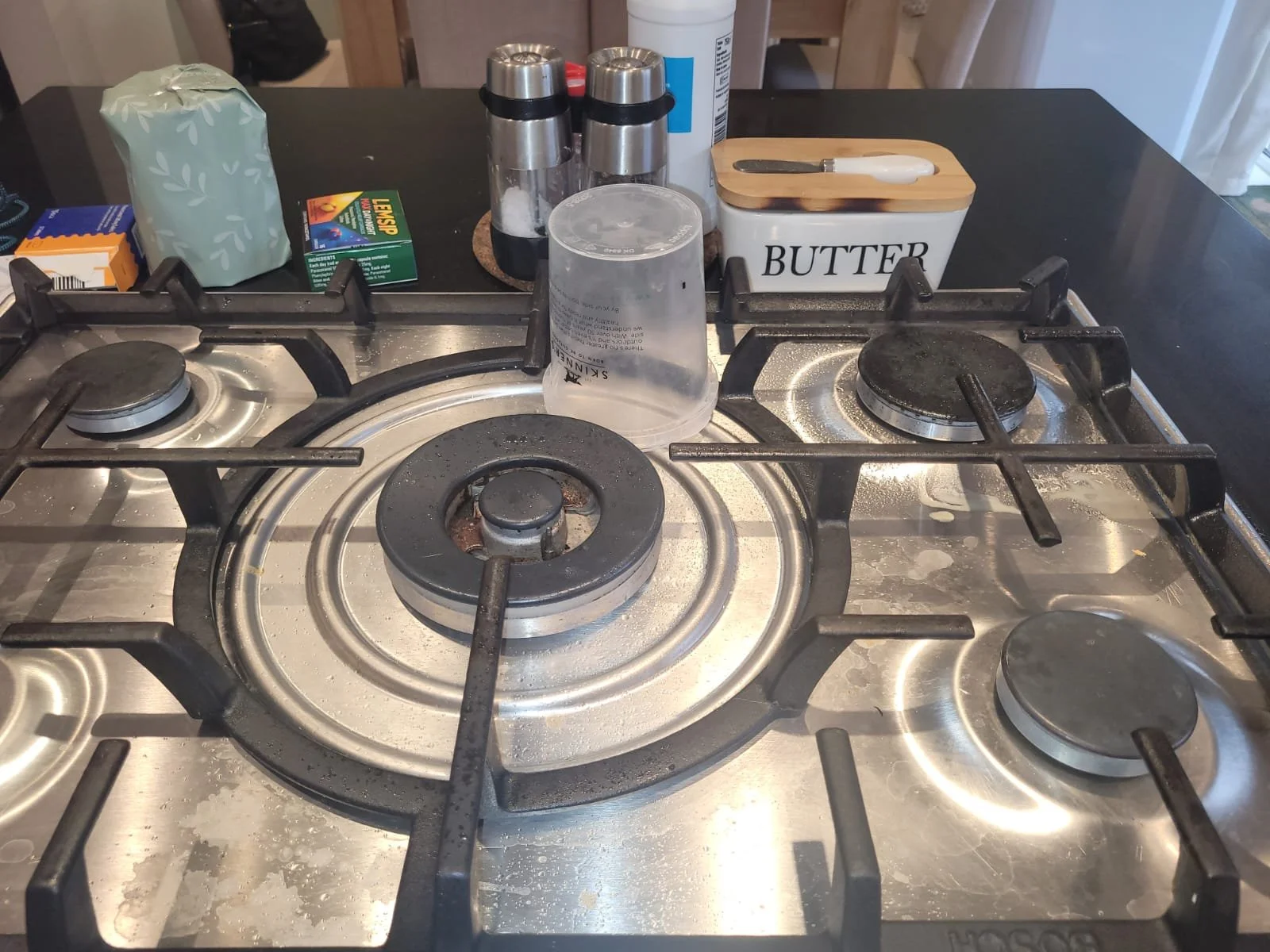 Stovetop with four burners, some with blackened surfaces, and various kitchen items in the background including a butter container, salt and pepper shakers, a tissue box, a cup, and small boxes.