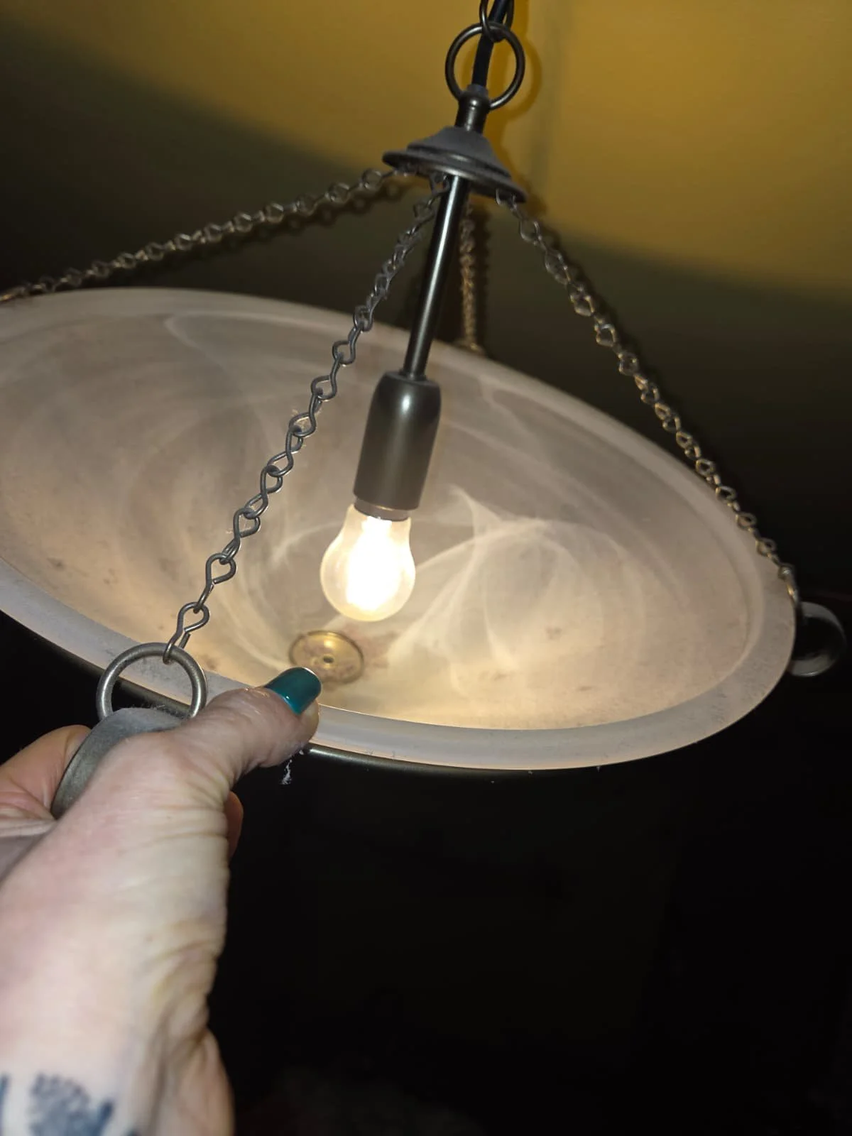 Close-up of a hanging light fixture with a chain, exposed light bulb, and a person's hand holding a small metallic object near the fixture.