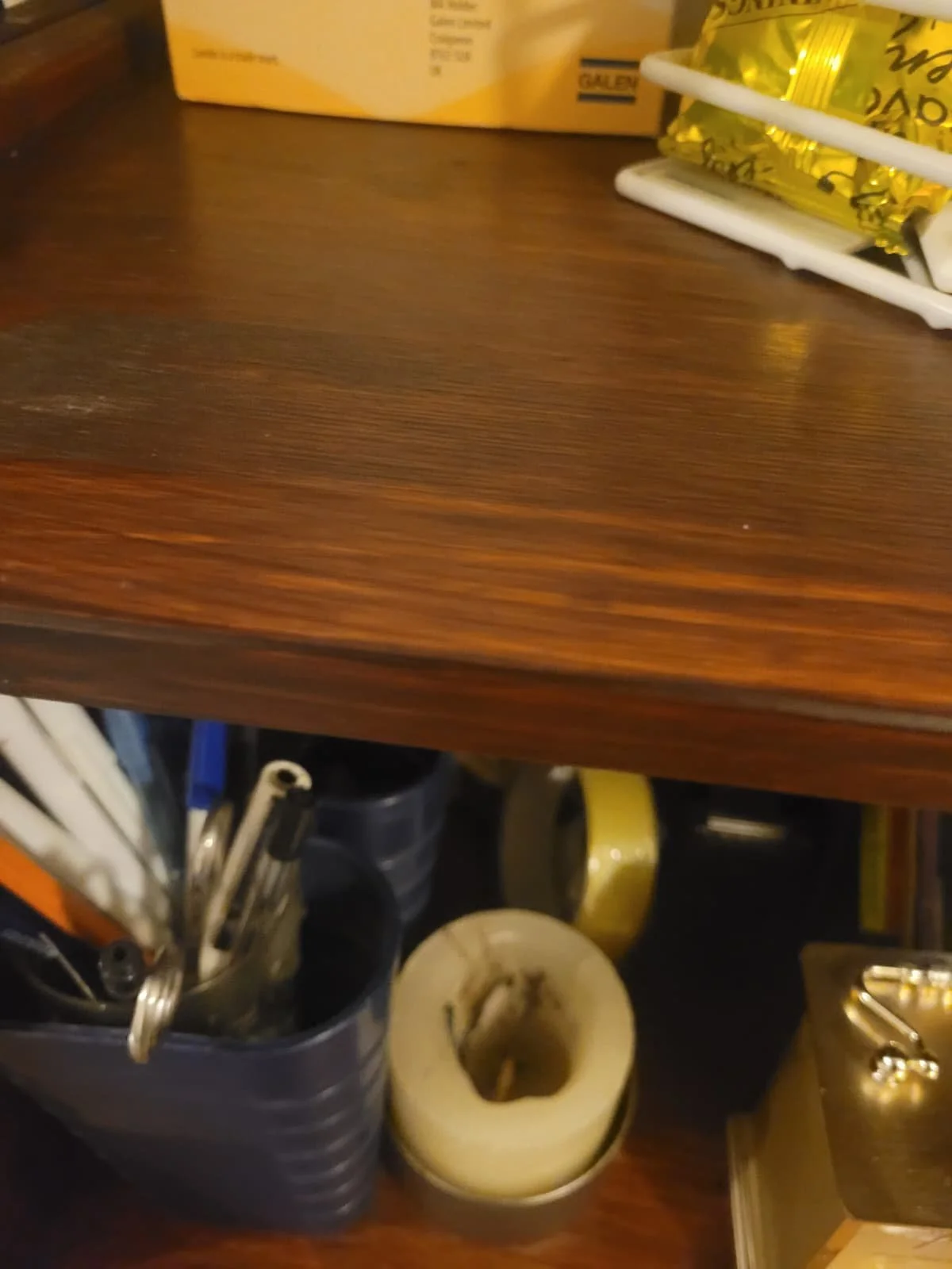 Part of a wooden desk with a container holding pens and a roll of tape underneath, with a yellow box and some yellow wrapped items on the desk.