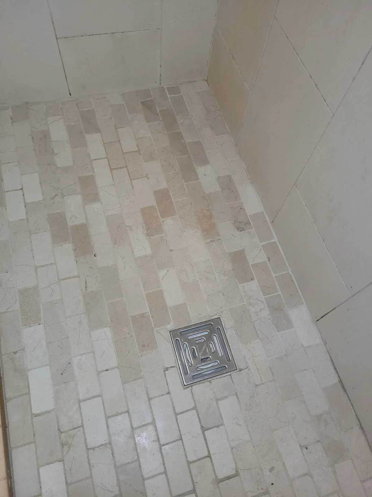 Close-up of a bathroom shower floor with beige mosaic tiles and a square metal drain.