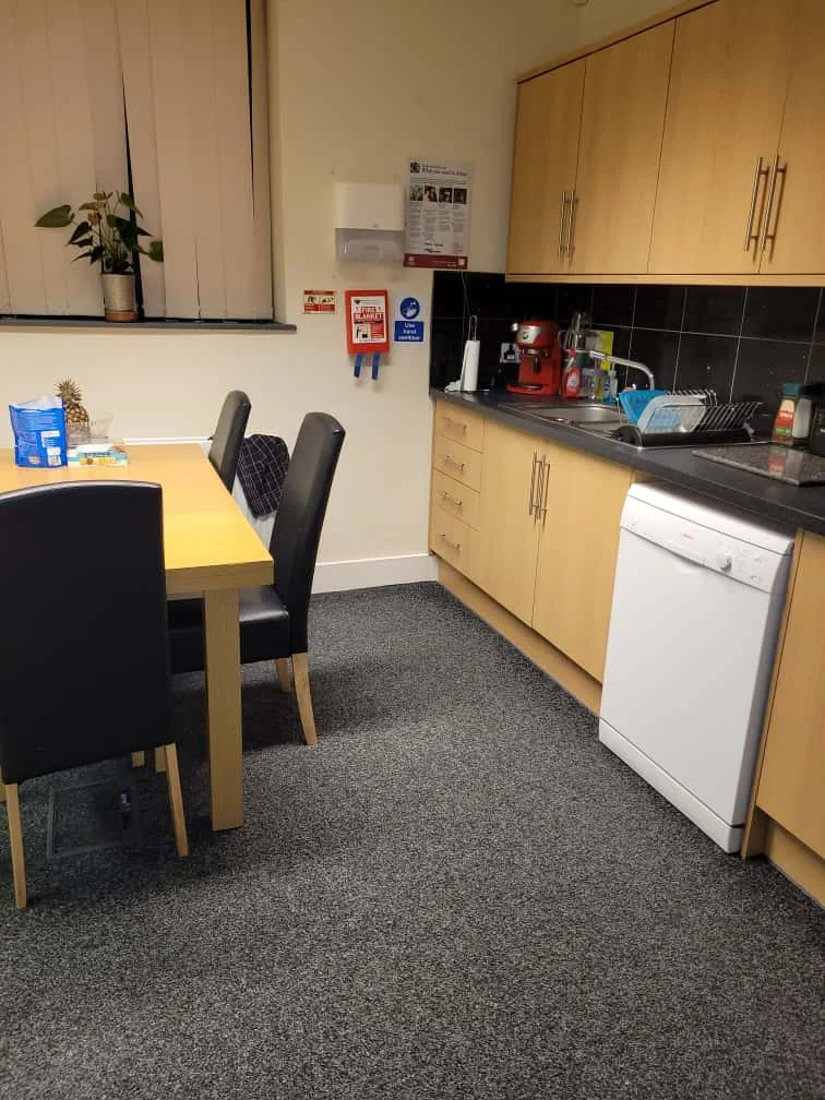 A small kitchen with a wooden table, four black chairs, a window with white blinds, a potted plant, and cabinets. The countertop has a dish rack, a coffee machine, and various kitchen items.