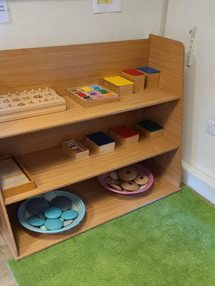 Wooden shelving unit with educational toys and materials, including colorful discussion stones, wooden blocks, and a tray of circular stones, set on a green carpet.