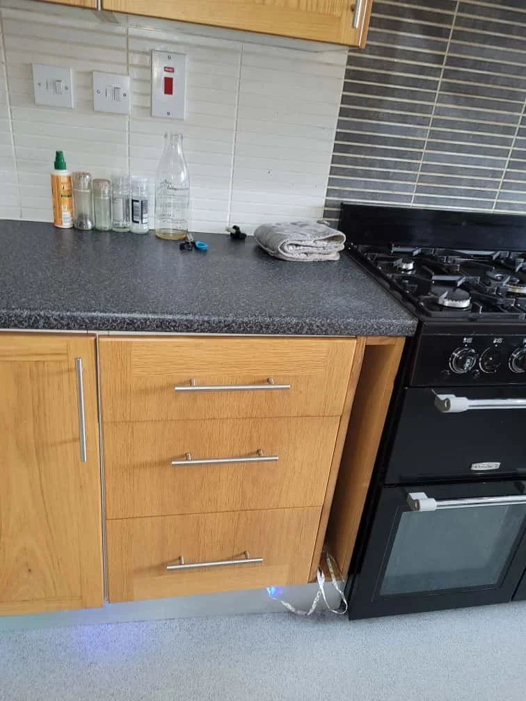 Kitchen countertop with glass bottles, cleaning spray, folded towel, and small objects next to a stove and wooden cabinets.