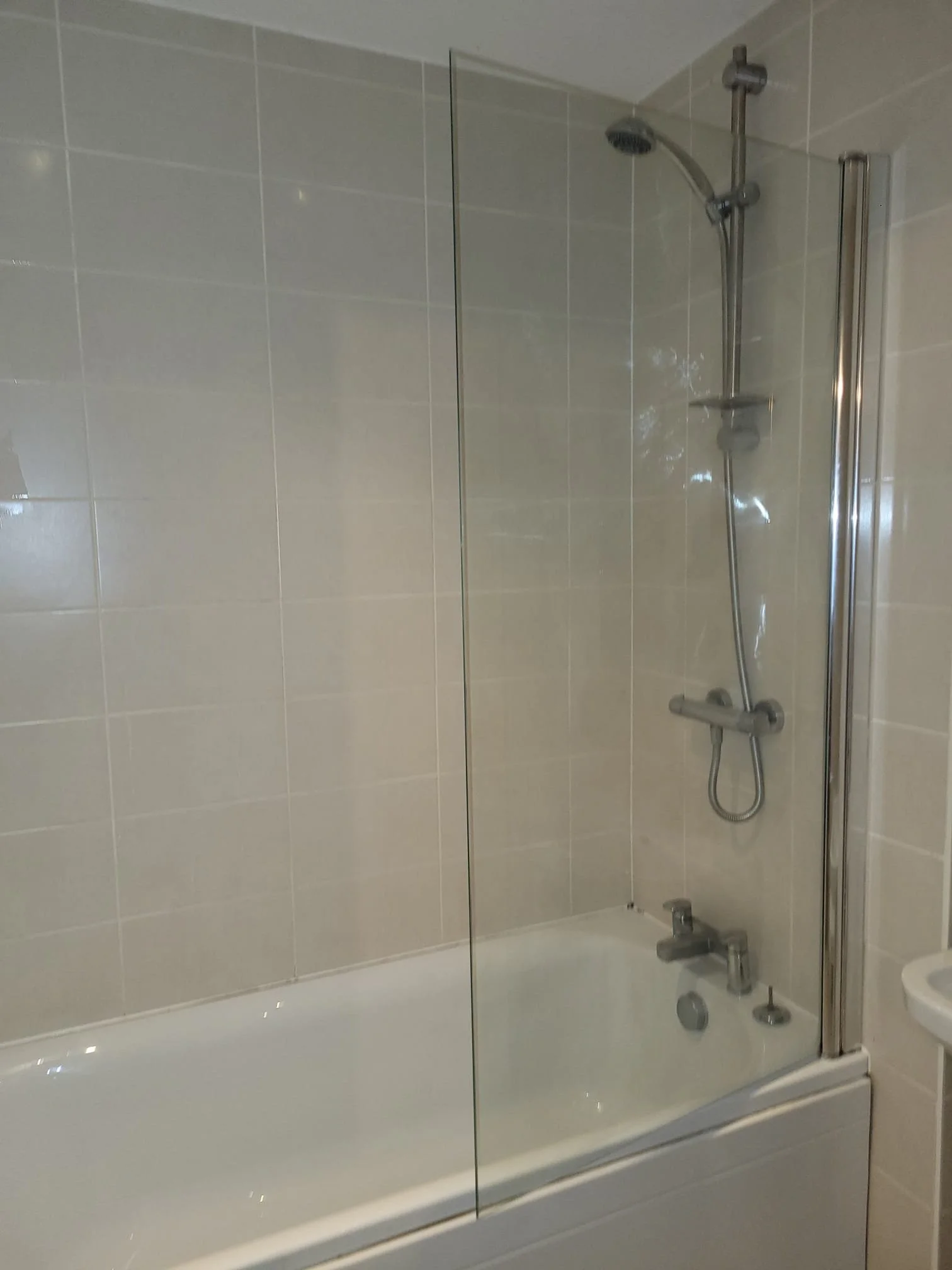 A bathroom with a bathtub and showerhead, enclosed with clear glass panels, tiled walls, and a small portion of a white sink visible on the right side