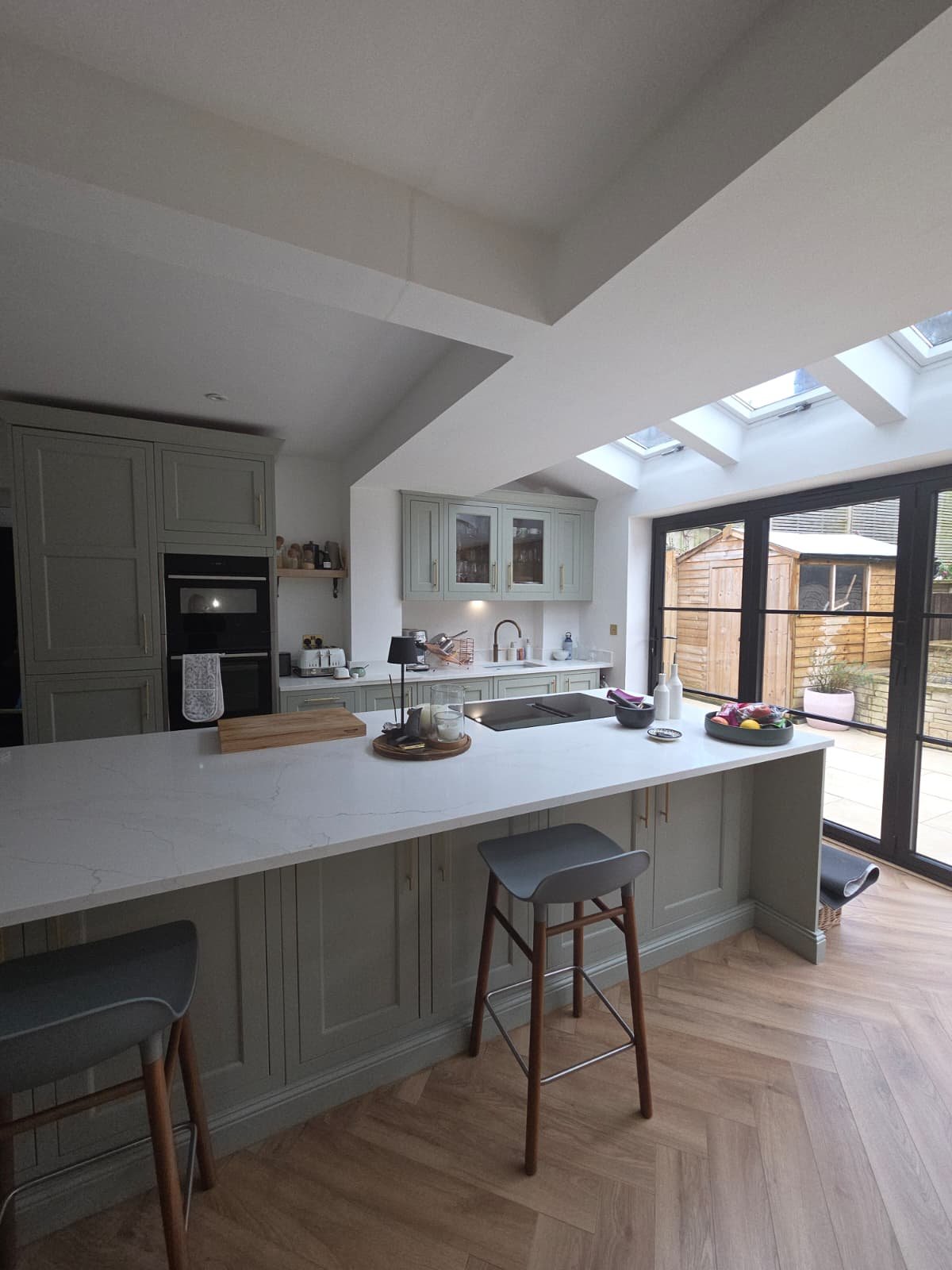 Bright kitchen with light green cabinets, a large white marble countertop island, and wooden bar stools. Skylights and large glass sliding doors lead to an outdoor patio.
