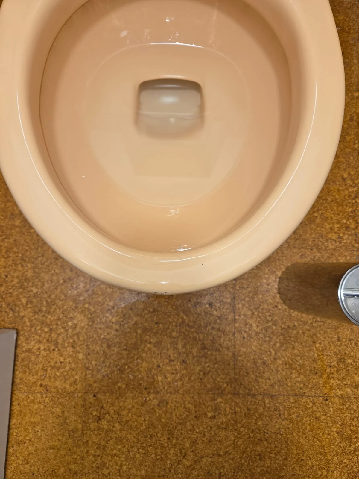 Top-down view of a beige public restroom toilet with the lid up, located on a brown speckled floor, with a metal trash can nearby.