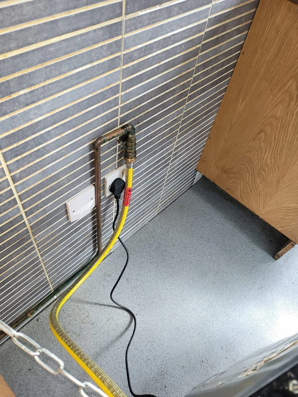 A utility area with pipe, electrical outlet, cord, and ventilation duct on a tiled wall and gray floor.