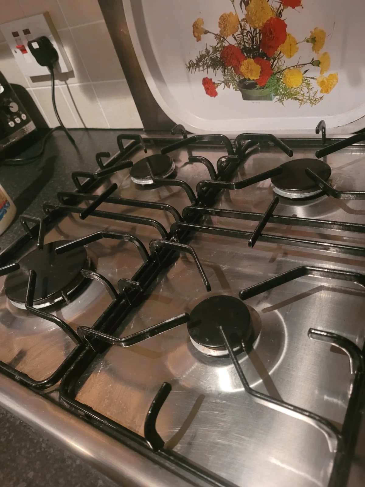 Close-up of a stainless steel gas stovetop with four burners and black cast iron grates, with a decorative flower scene on a white panel in the background.