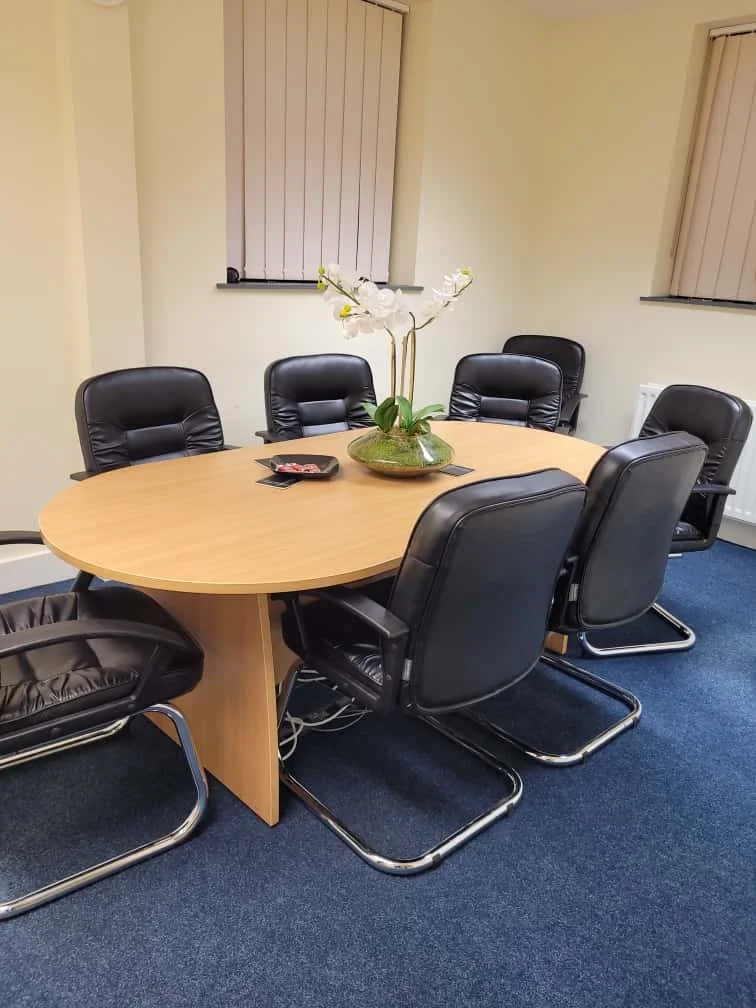 Conference room with a large oval wooden table, six black leather chairs, a potted orchid plant in the center, two windows with blinds, and a blue carpet.