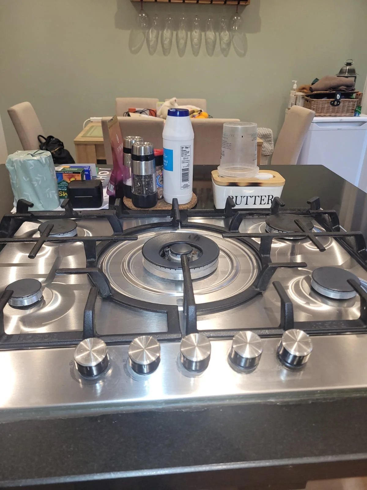 Close-up of a stainless steel gas stove with five burners in a kitchen. In the background, there is a dining area with chairs, a table, and various household items.
