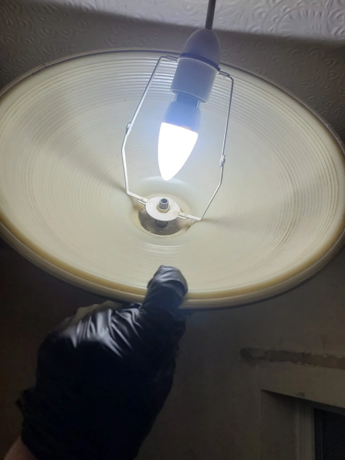 Person wearing a black glove pointing at the inside of a large ceiling lamp with the light bulb turned on.