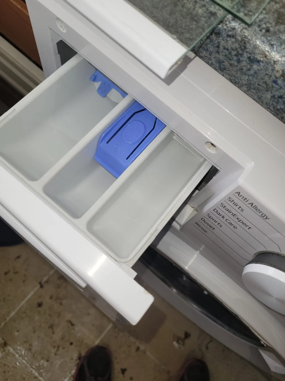 Open laundry washing machine drawer with a blue detergent compartment, with an allergy warning sticker on the side.