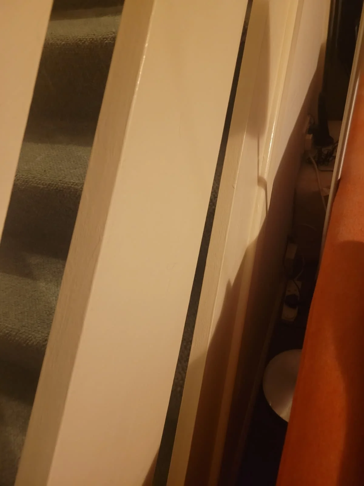 A staircase with gray carpeted steps next to a white wall and a door frame with a small gap revealing a glimpse of a cluttered space with wires and objects.