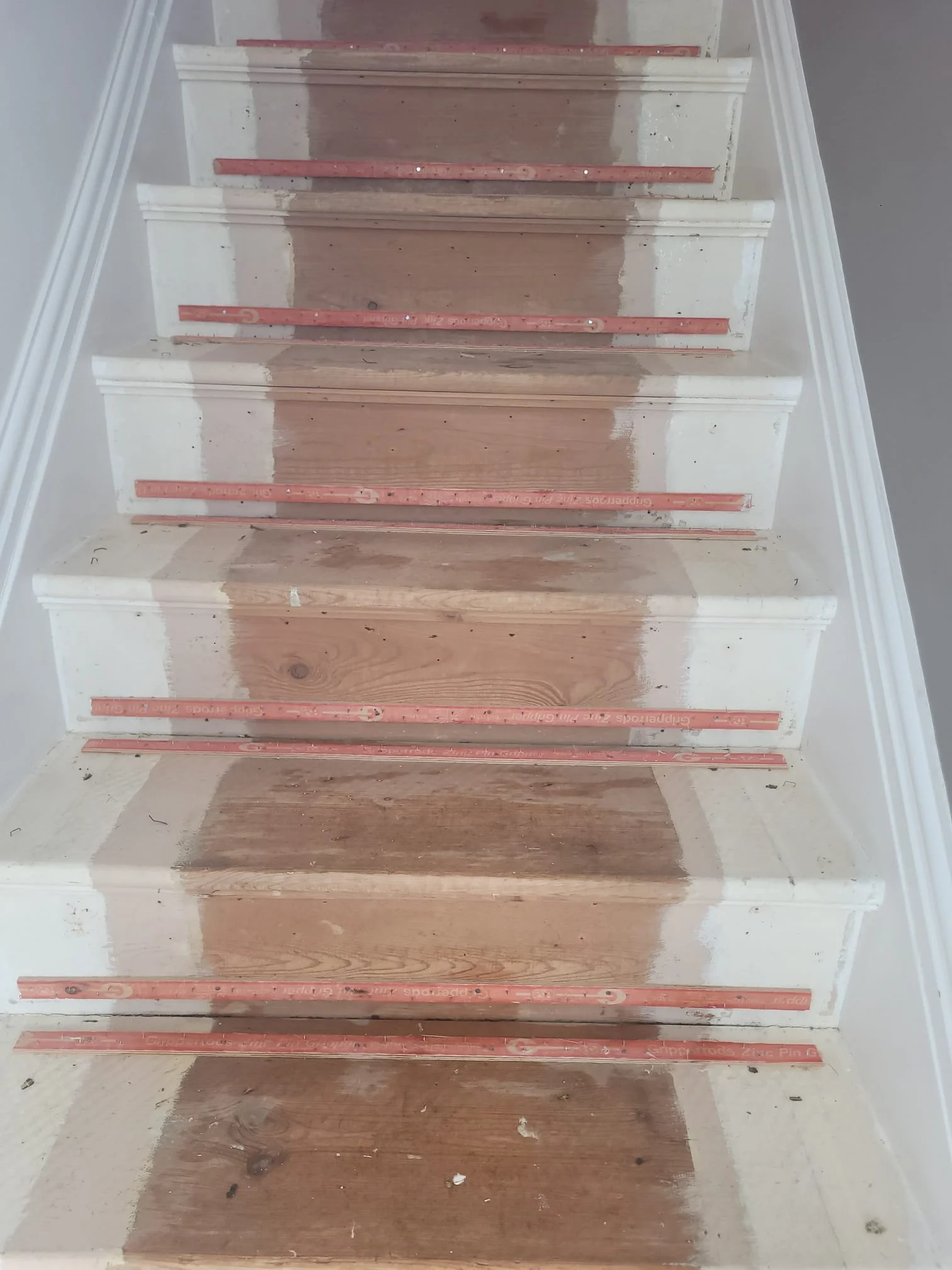 Staircase under renovation showing partially painted white risers, unpainted wooden treads, and red construction leveling tools placed along each step.