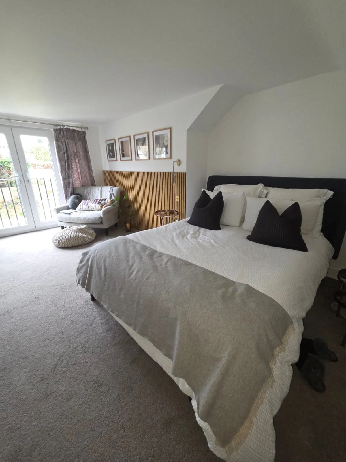 Bedroom with a large bed, dark headboard, white bedding, and black pillows. Small sitting area with a light-colored armchair, patterned pillow, and a round woven ottoman. French doors with glass panels and curtains leading to a balcony. Wood-paneled wall with framed pictures, and a small table with a plant.