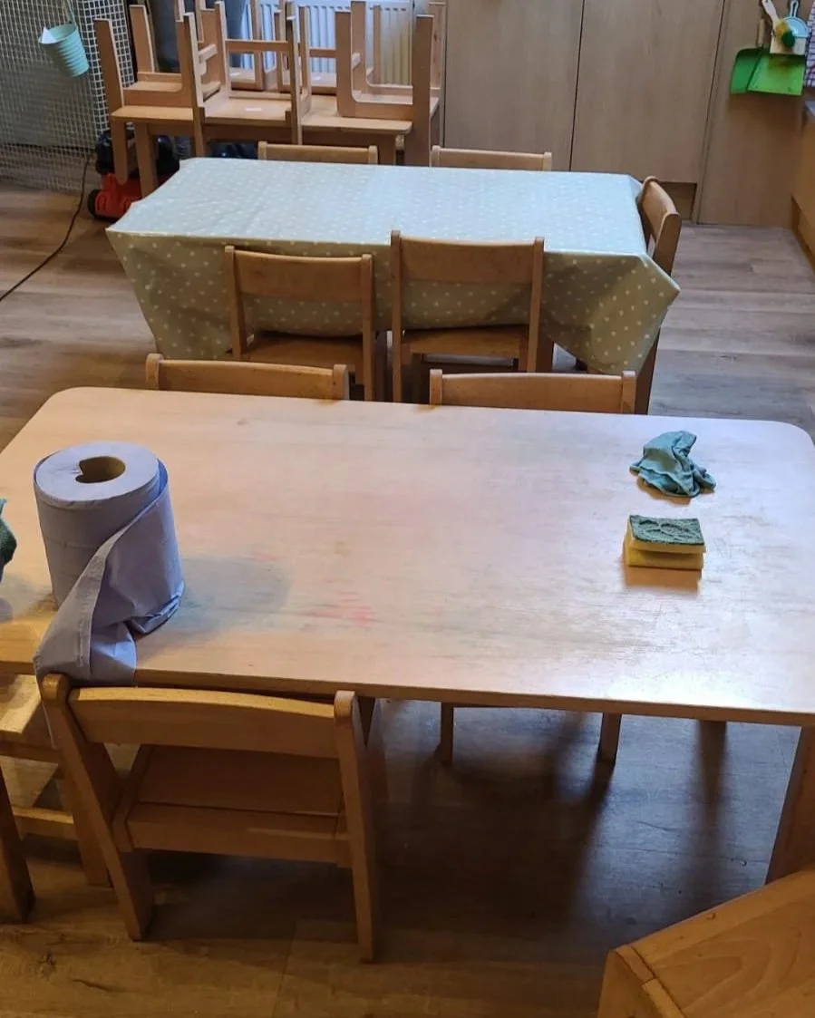 A wooden table with cleaning supplies and cloths, set in a room with multiple smaller tables with polka-dot tablecloths and chairs, likely a classroom or daycare setting.