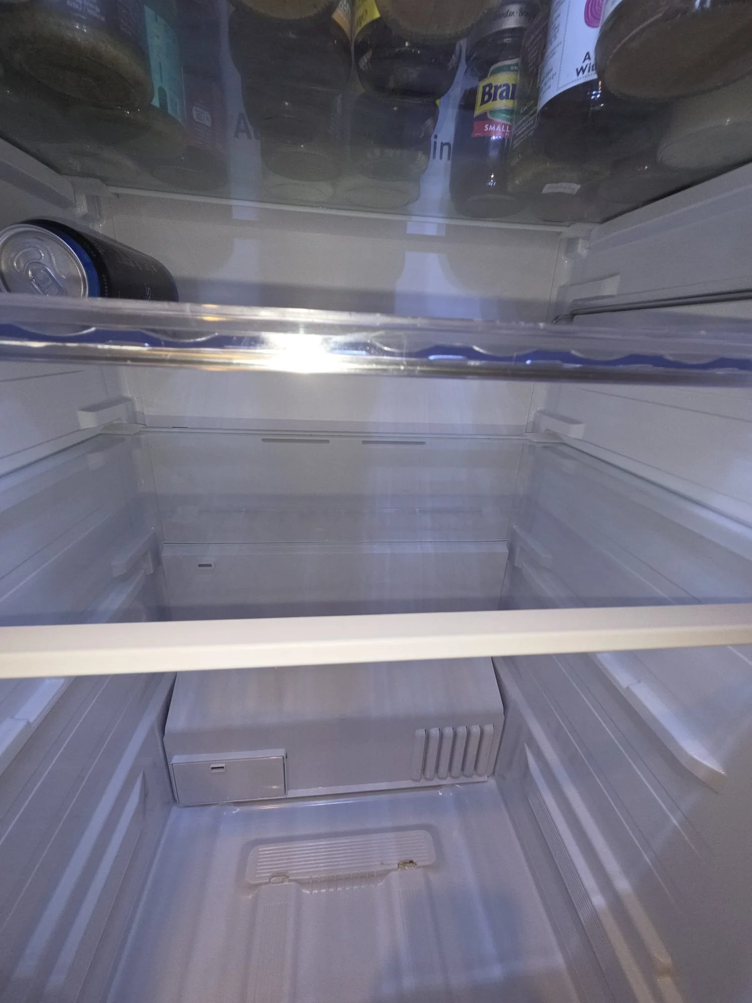Empty refrigerator with a few bottled drinks on the top shelf and a single soda can inside.