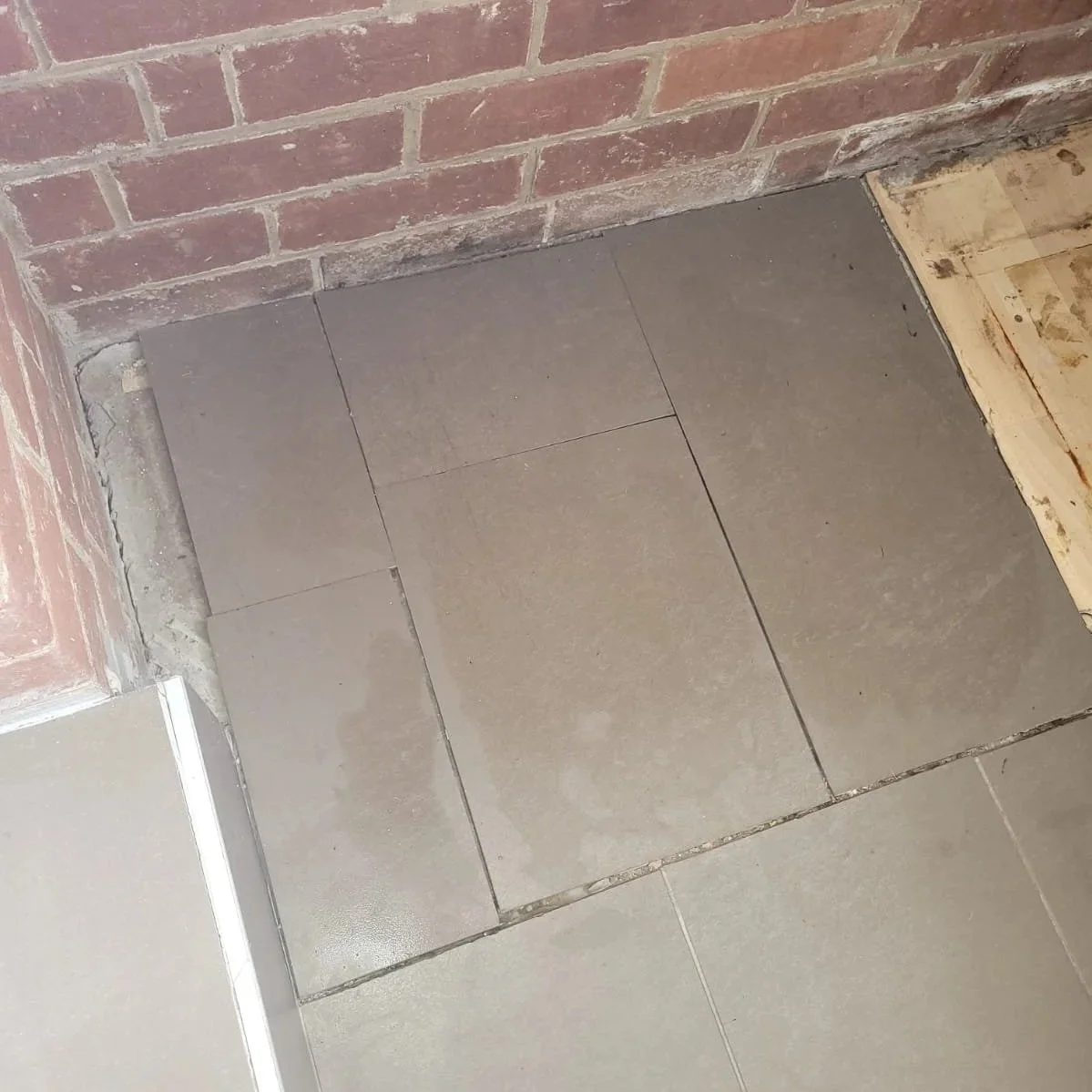 Close-up of a corner with gray tiles on the floor, brick wall, and new wooden floorboard.