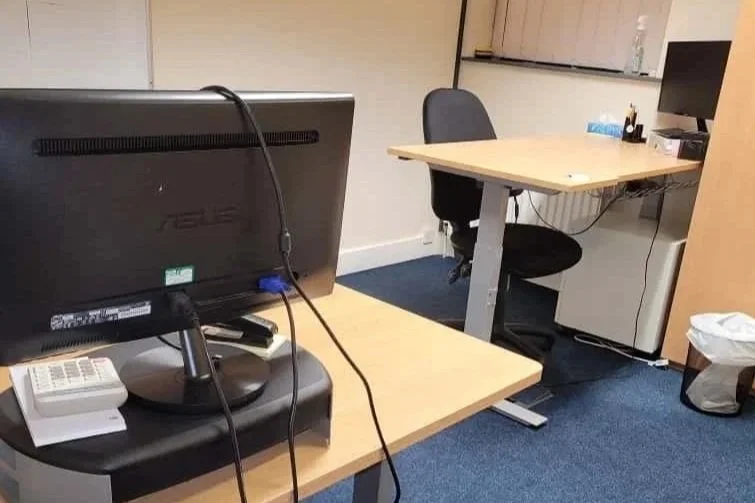 An office desk with a computer monitor, a landline phone, and a black chair, next to a smaller desk with a computer and office supplies, in a room with blue carpet.
