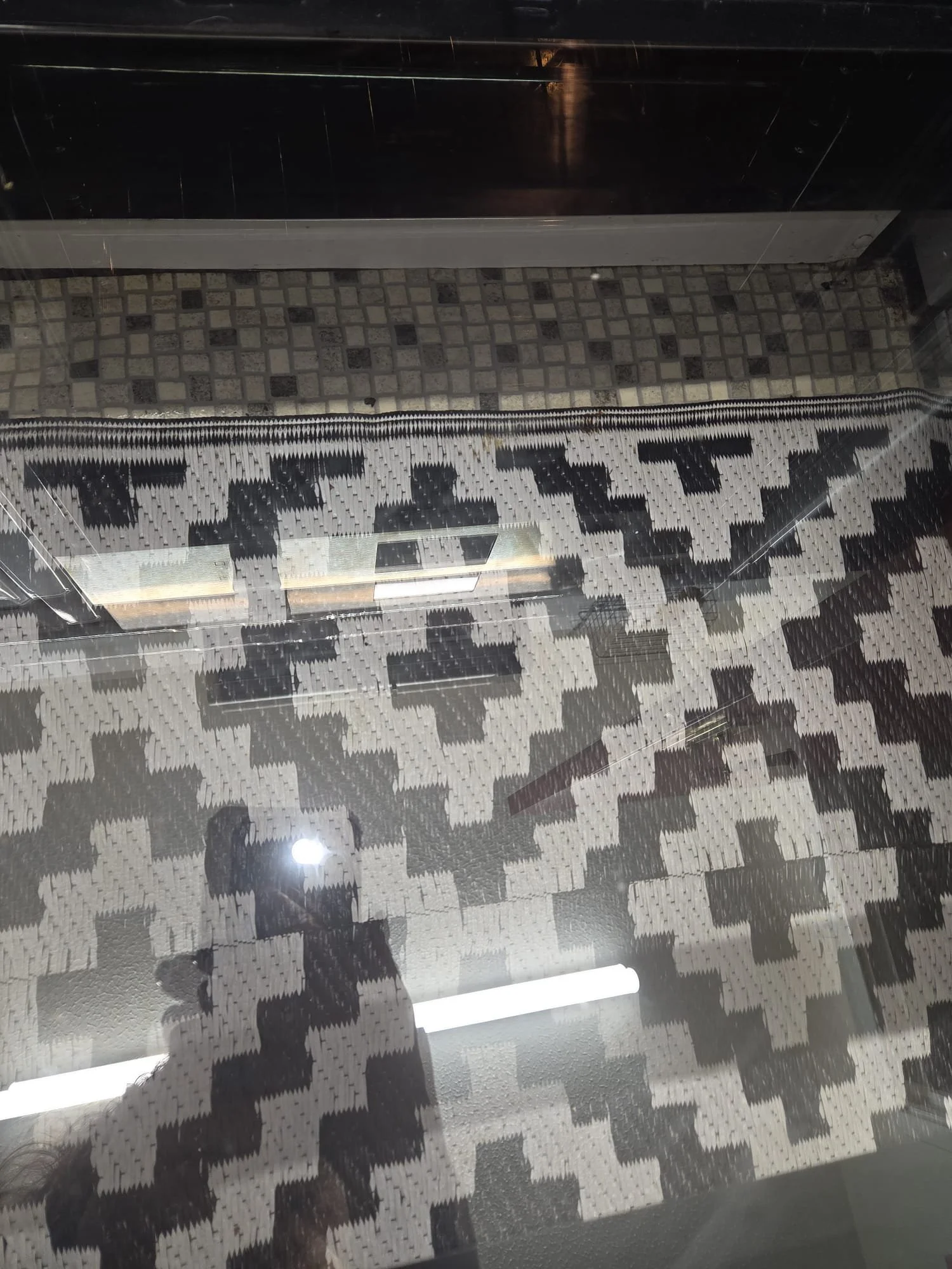 A glass display case with a patterned rug beneath it, reflecting ceiling lights and part of a person's shadow.