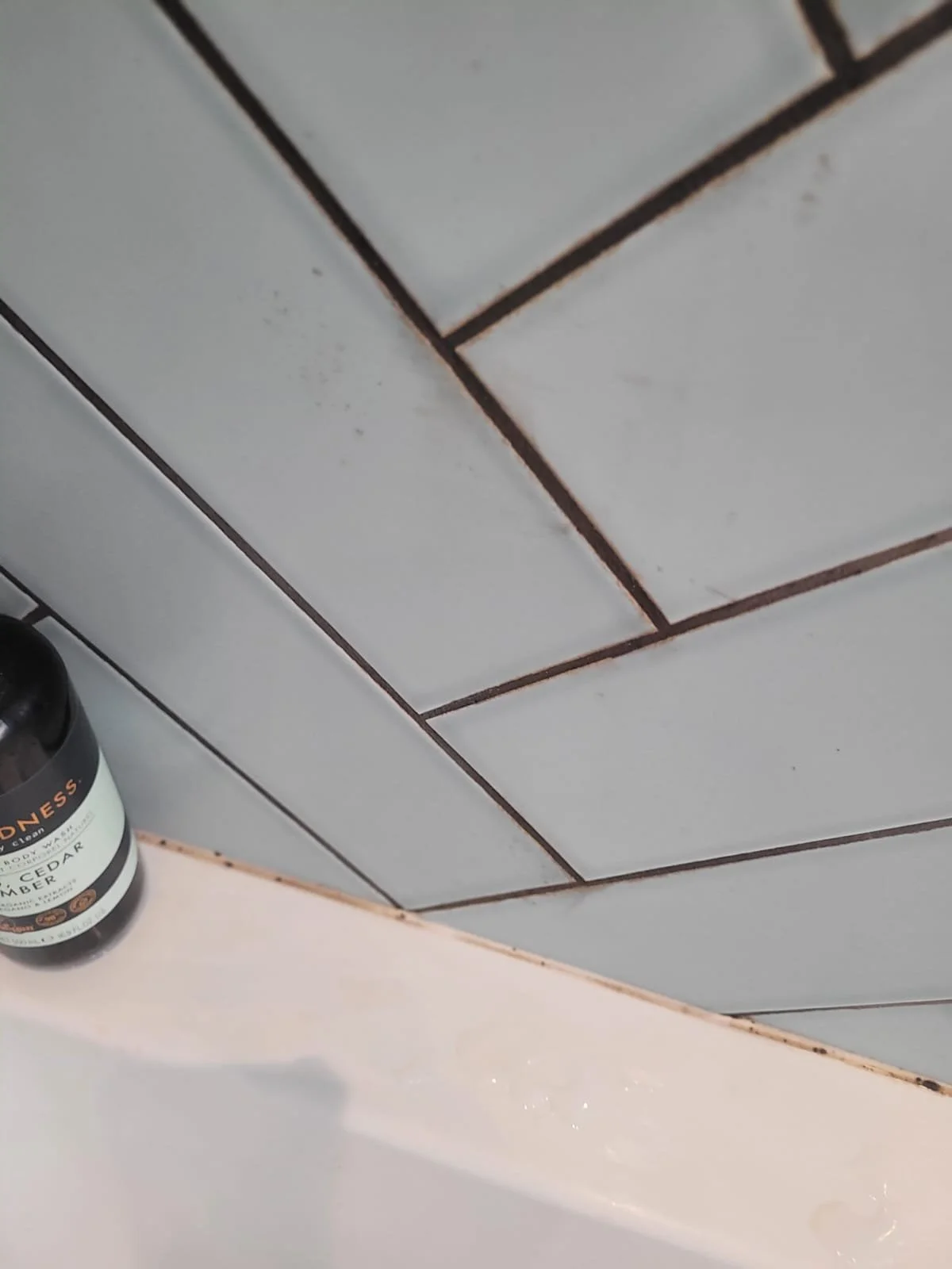 Close-up of tiled bathroom wall and bathtub edge with a bottle of body wash or shampoo on the bathtub rim.