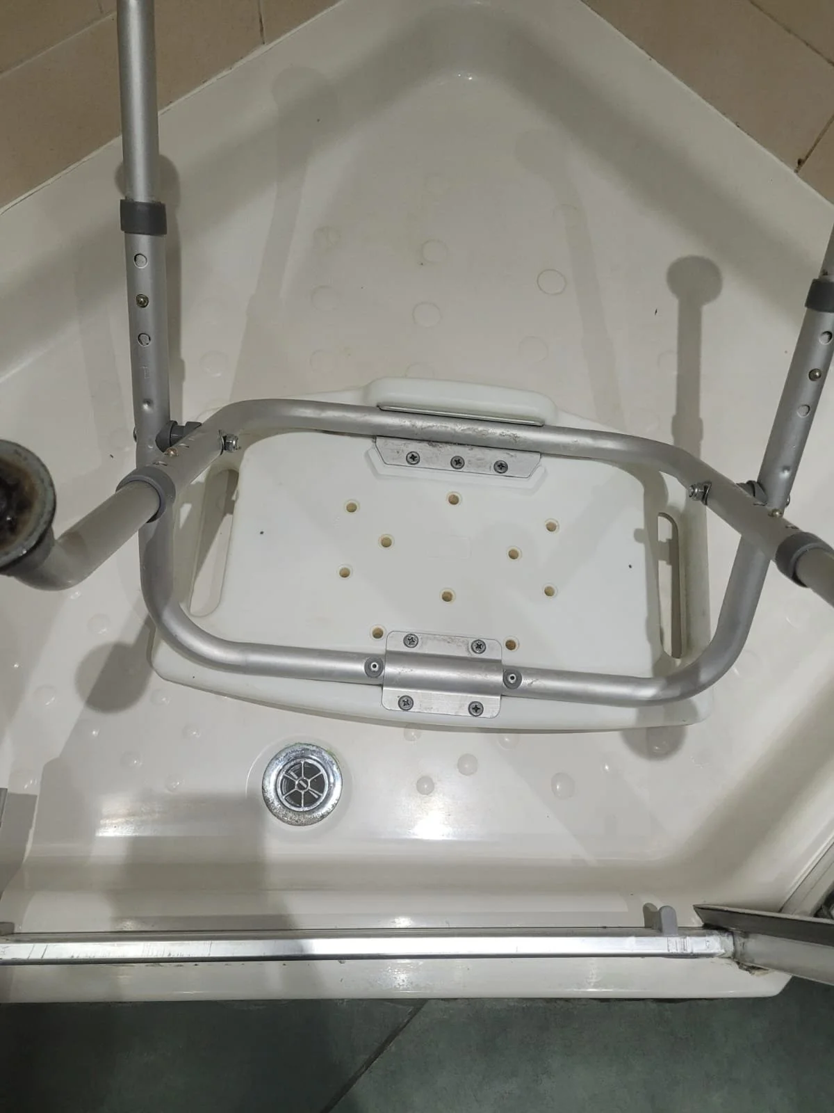 A shower chair with a metal frame placed inside a shower stall with a drain on the floor.