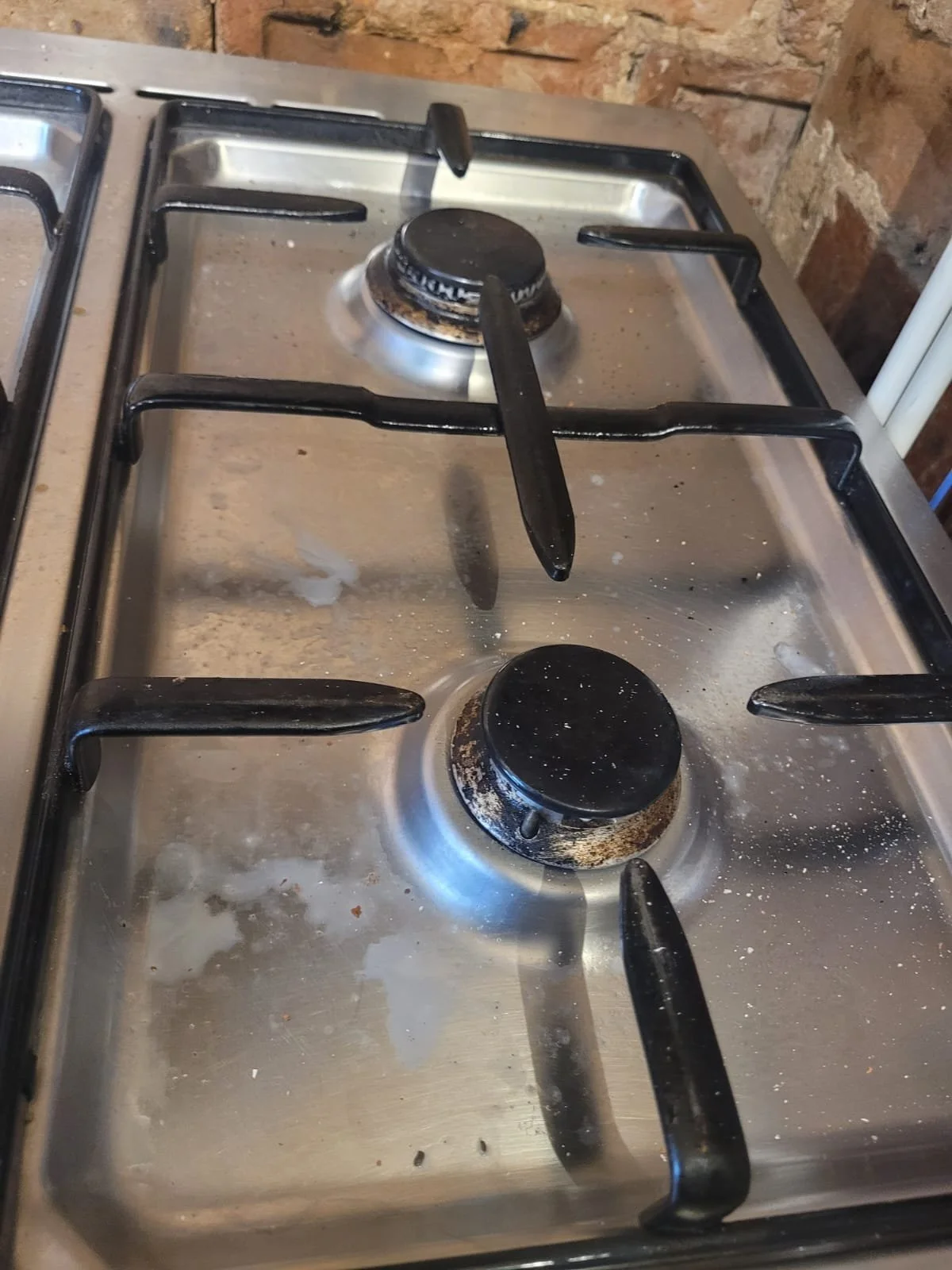 Dirty, stained stovetop with two burners, one with a rusted burner cap, and black grates, in a kitchen with brick walls.