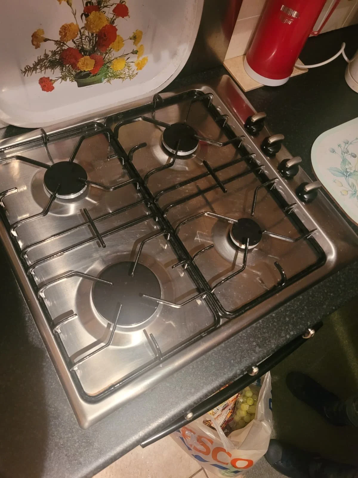 A stainless steel gas stove with four burners on a kitchen countertop. There are floral decorative items, a red container, and a plastic bag with groceries nearby.