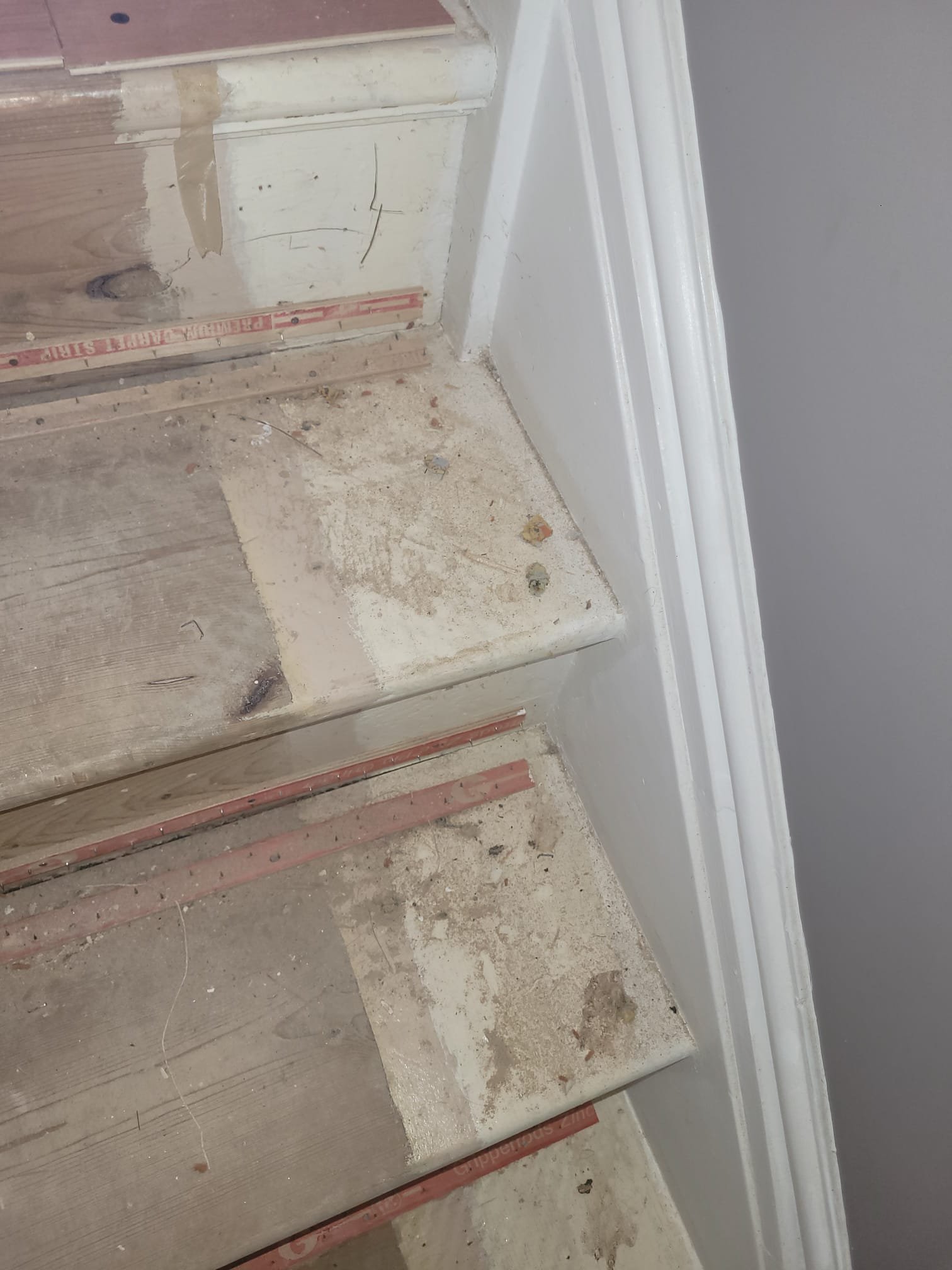 Close-up of a section of a staircase under construction, showing wooden stairs, dust, dirt, and part of a gray painted wall with white trim.