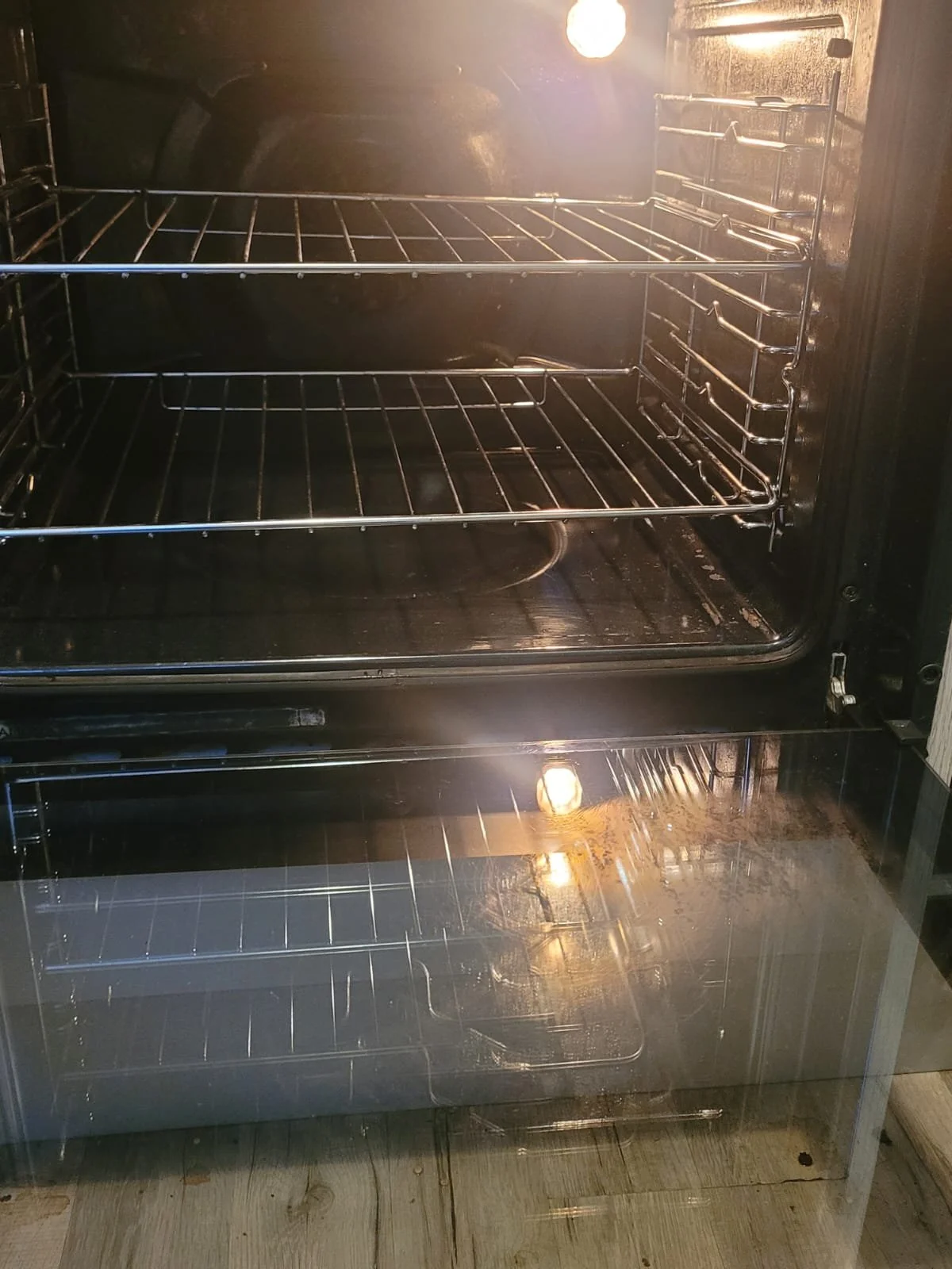 Empty oven with glass door, multiple wire racks, lit interior light, and wooden floor reflected in the glass.