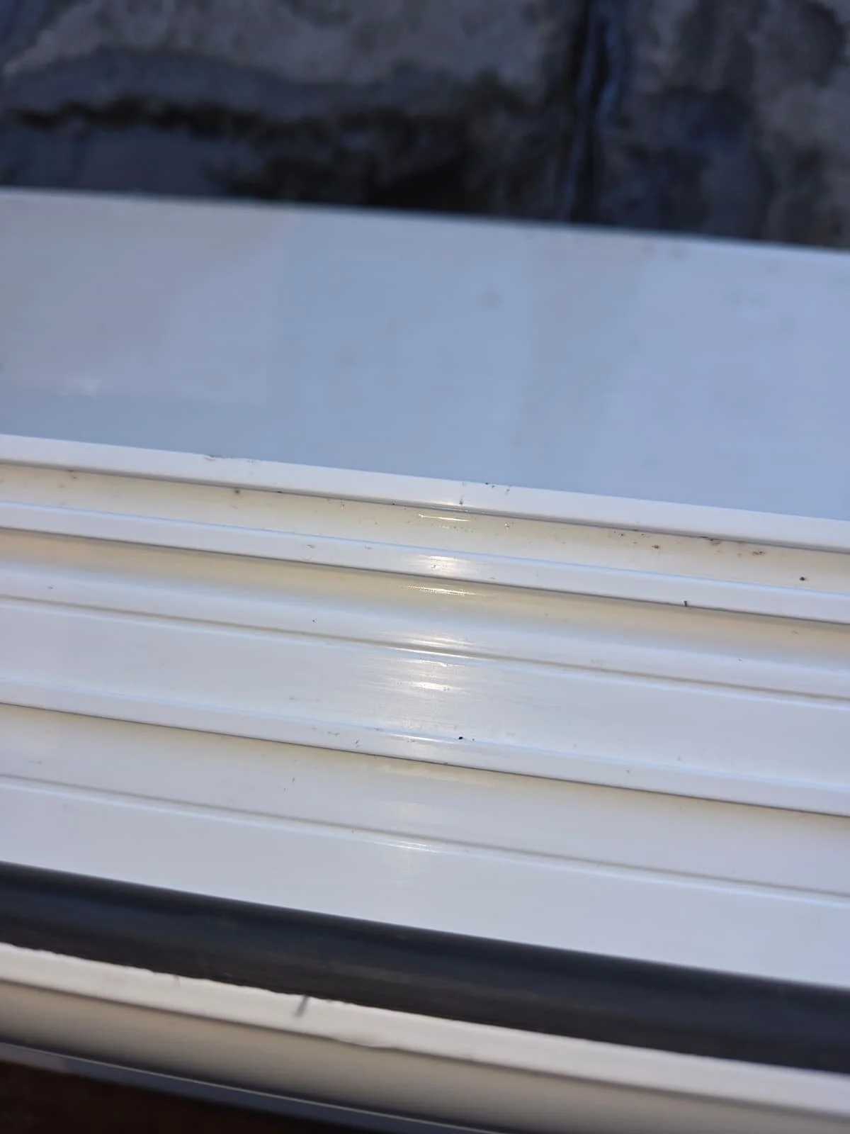 Close-up of a white plastic or metal window or door frame with multiple layers and a black rubber seal at the bottom, with a dark stone or concrete surface in the background.