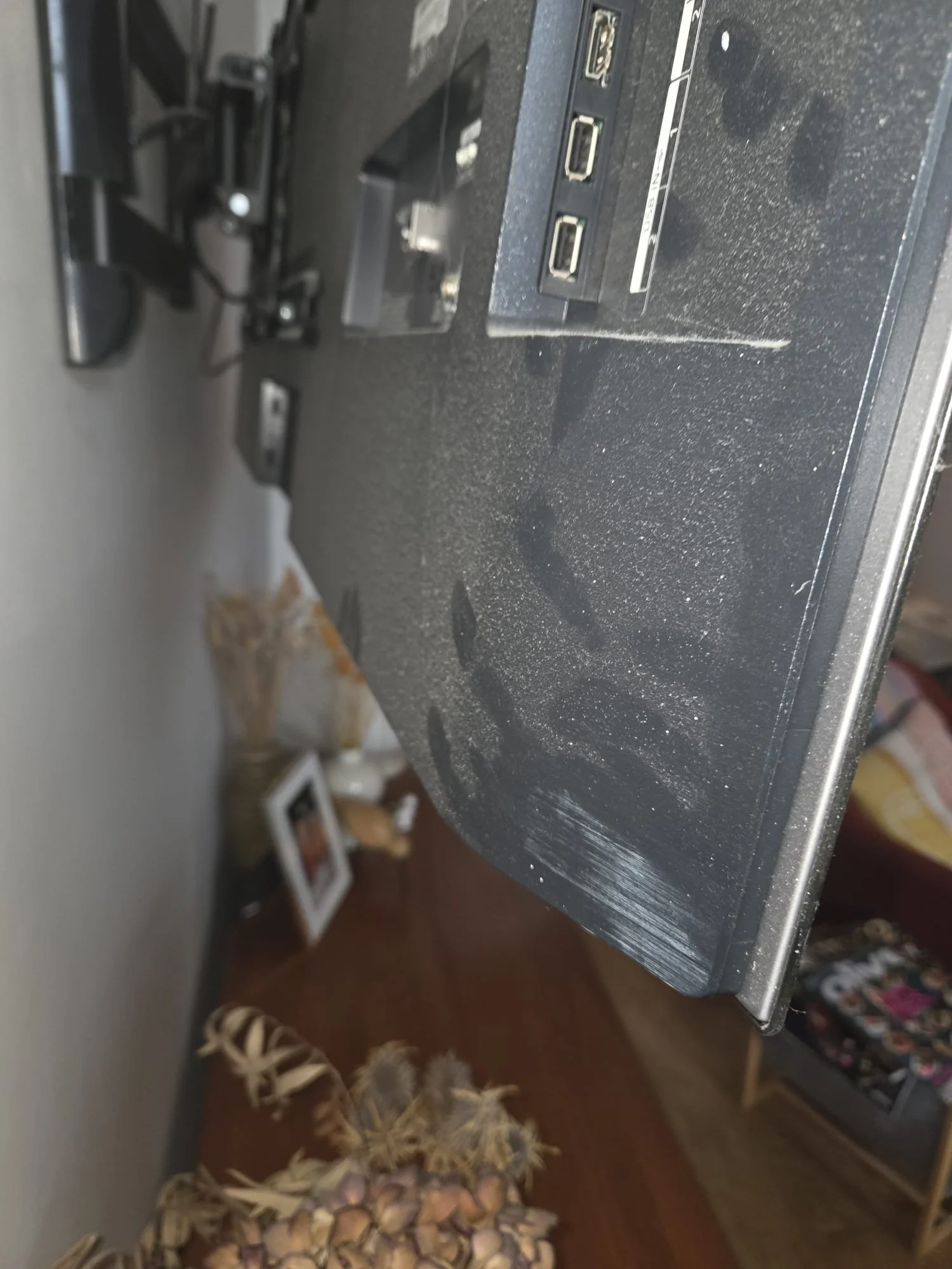 Close-up of a black electronic device with three USB ports on a desk, with a dusty surface and visible scratches. In the background, there is a person with long blonde hair, a framed photo, and a container of garlic cloves on a wooden table.