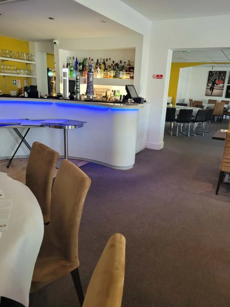 Empty restaurant bar area with a curved counter, alcohol bottles on shelves, and beige chairs around tables. Bright lighting and colorful walls create a modern atmosphere.