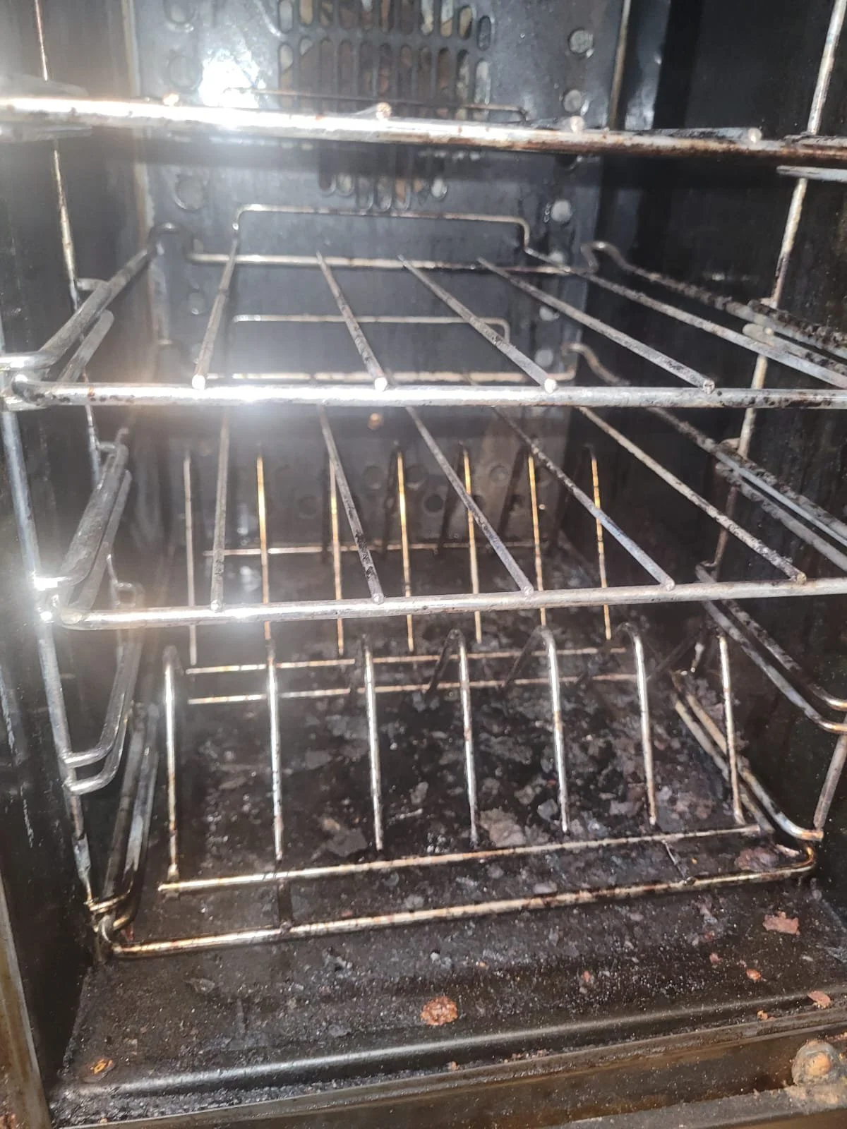 Inside of a dirty oven with blackened surfaces, racks, and debris.