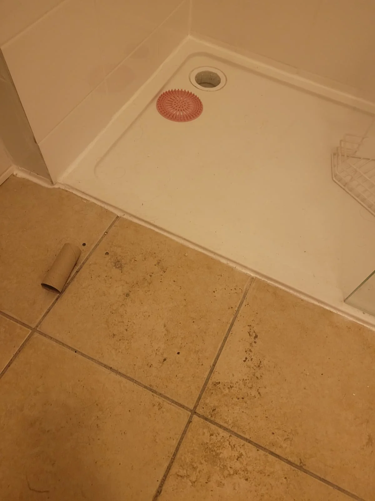 Bathroom shower with a drain cover, a hole in the shower floor, a piece of cardboard on the floor, and tiled flooring.
