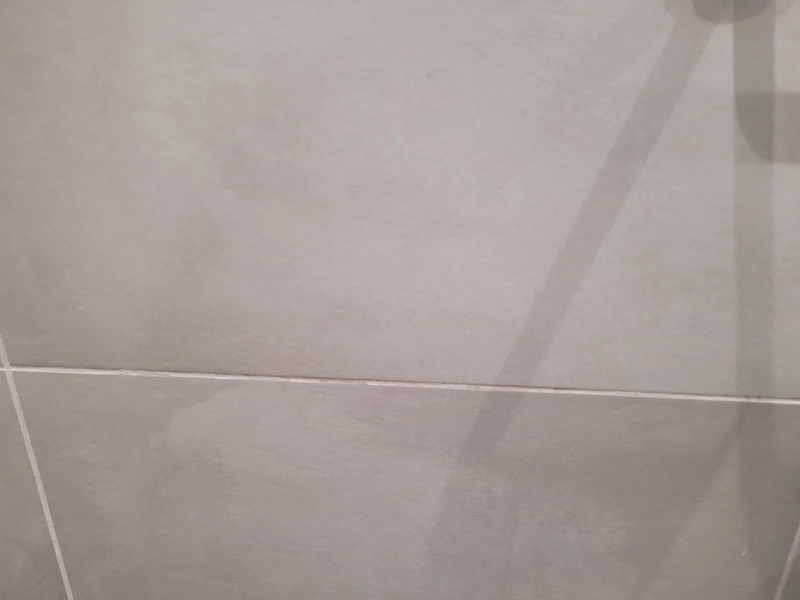 A close-up of beige/gray tiled ceiling with a visible grout line and a ceiling fan shadow.