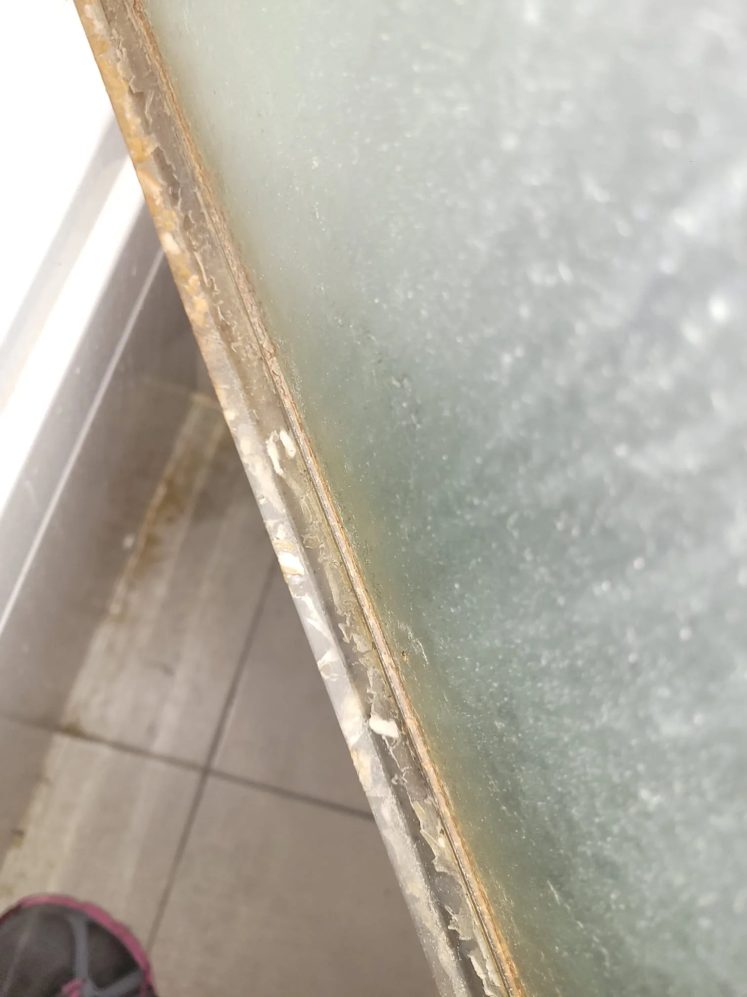 Close-up of a glass panel with rust and corrosion along the metal frame, positioned at an angle in a room with tiled flooring.