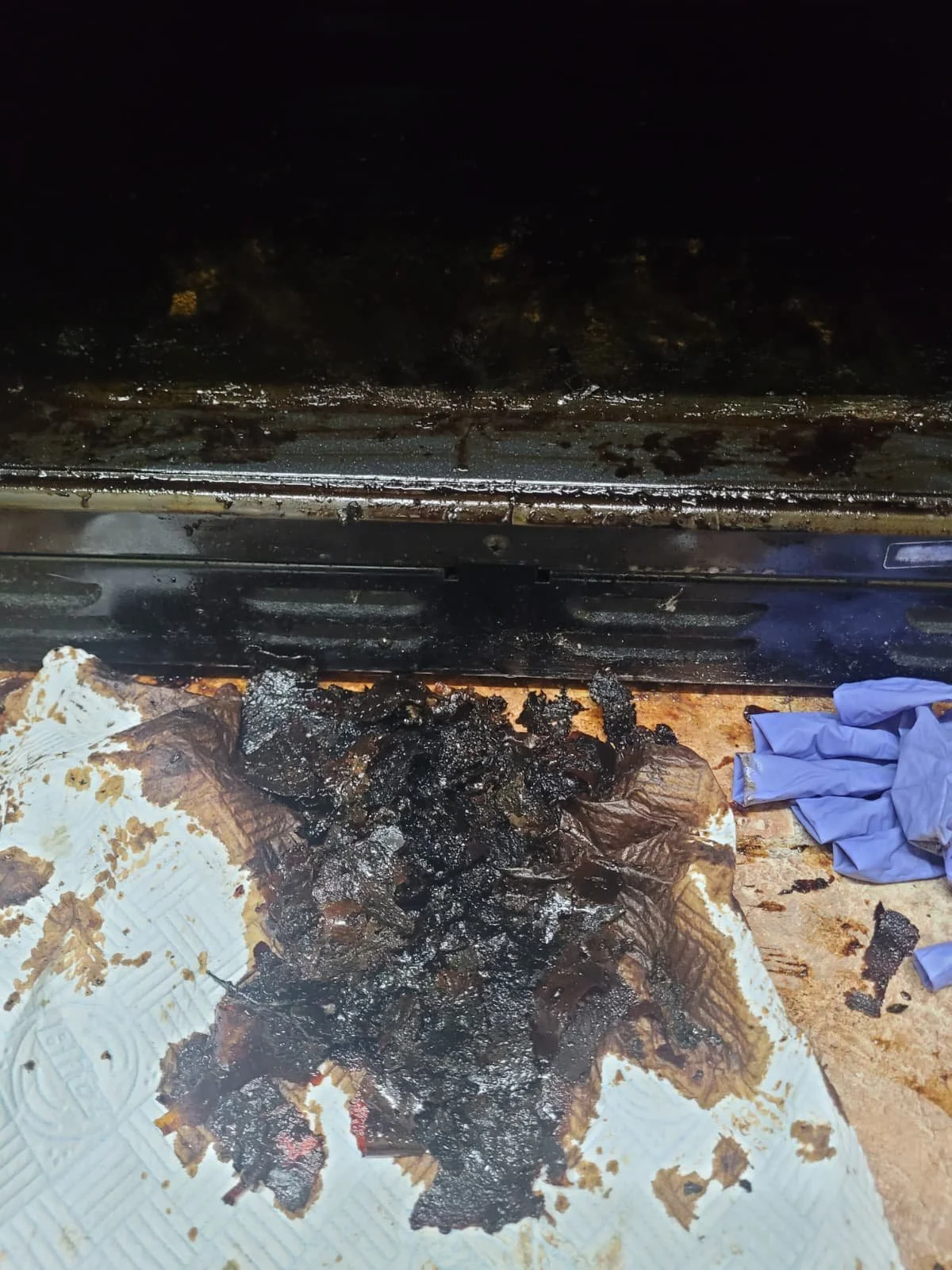 Charred and burnt food residue on a paper towel, with a pile of crumpled blue gloves nearby and an oven or appliance in the background.