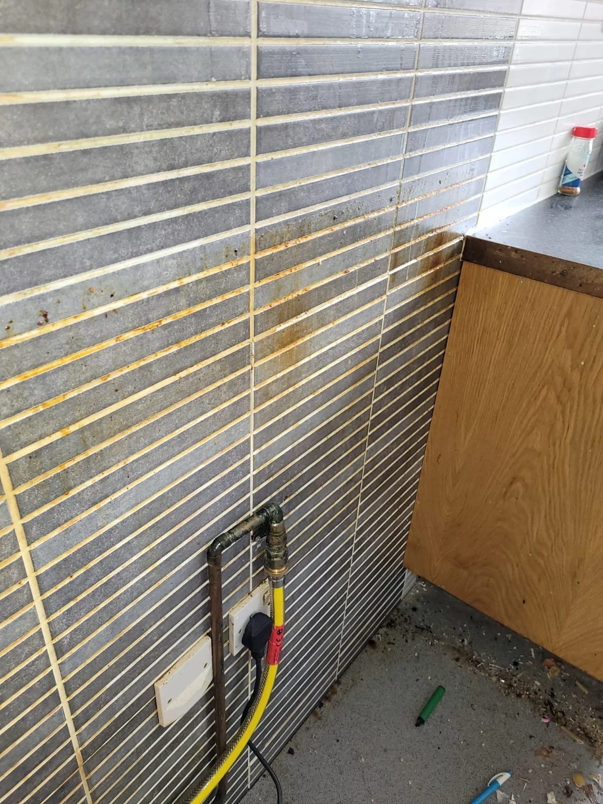 A tiled wall with gray and white tiles and yellow grout, showing rusty stains near the corner. A gas pipe with a yellow hose is connected to a wall outlet, with some debris and a few scattered objects on the floor.